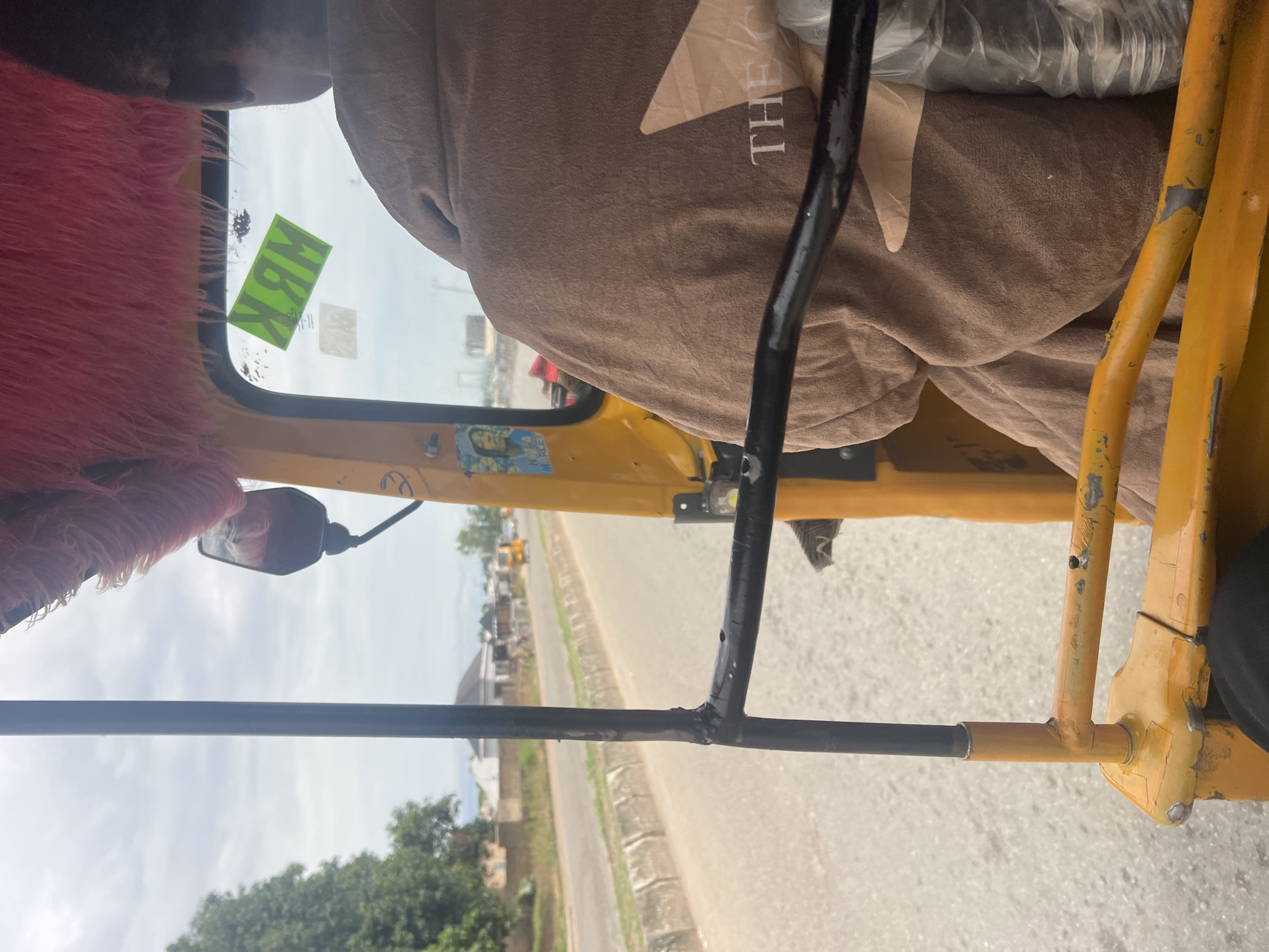 The image provides an interior view from the passenger seat of a yellow motorized rickshaw, likely in Jalingo, Nigeria. The rickshaw's interior is decorated with a vibrant pink, fluffy material on the left side, possibly a seat cover or trim. On the windshield, a bright green sticker with black lettering is visible, which appears to read "NBK" when viewed from the driver's perspective, indicating it is placed for the driver to see. To the right of the sticker, another smaller, faded sticker or decal is present.
A brown fabric, possibly a blanket or a seat cover, is draped over a portion of the rickshaw's interior to the right. A faint, white stylized "THE" is visible on this fabric, suggesting it might be branded or personalized. A black metal bar, part of the rickshaw's structure, runs vertically through the center of the frame.
Through the windshield, a cloudy sky is visible, suggesting overcast weather. The scene outside depicts a road with a few buildings and trees lining its side. In the distance, other vehicles, possibly more rickshaws or cars, can be seen on the road, indicating a daytime activity. The overall impression is a snapshot of everyday travel within a Nigerian urban or semi-urban environment.