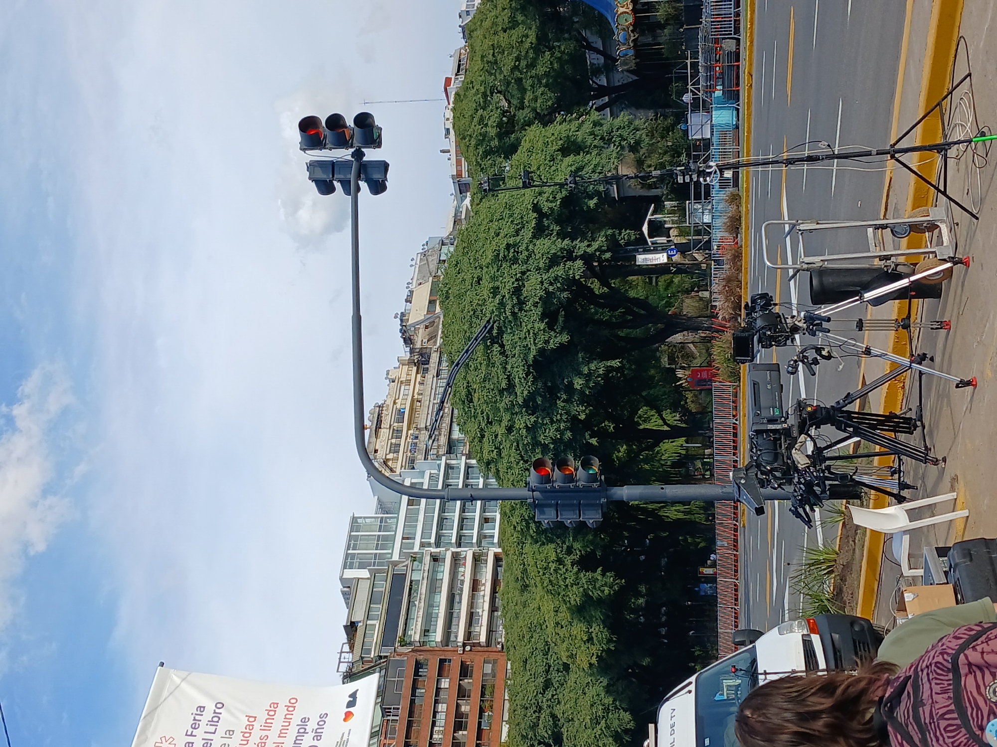 The image shows a city street scene in Buenos Aires, Argentina, likely during an event related to "La Feria del Libro de la Ciudad" (The City's Book Fair), judging from the banner on the left. There are traffic lights visible, with the top set displaying a red light and the bottom set showing a transition from red to green. The surroundings include buildings, trees, and street furniture. Several professional video cameras are set up on tripods along the side of the street, suggesting live broadcast coverage. The street markings and a white van with "IL DE TV" are visible. The presence of people is indicated, although their faces are not clearly visible. The weather appears to be clear with partly cloudy skies.