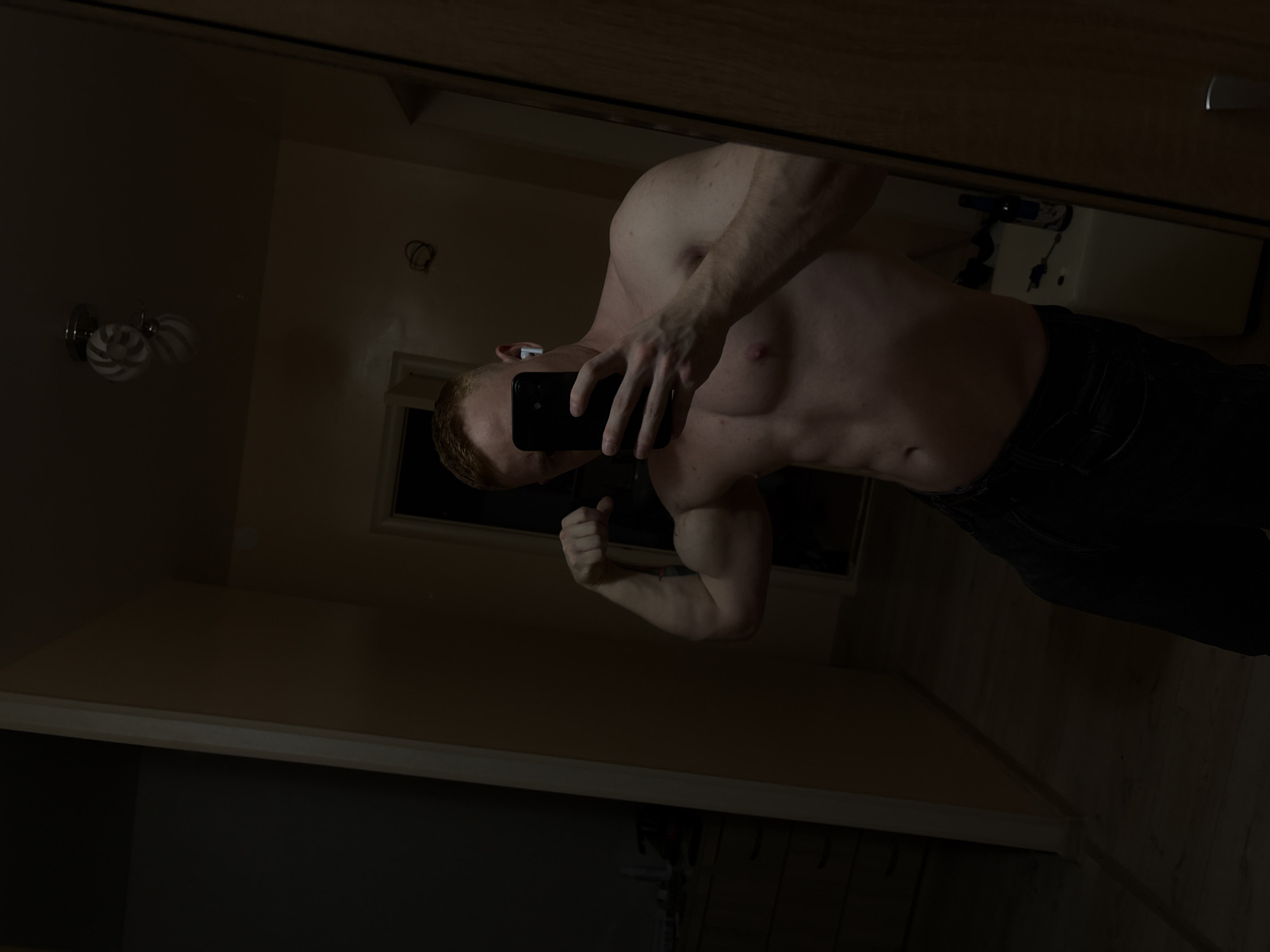 A shirtless man is visible in a mirror, flexing his bicep. He is holding a black smartphone and appears to be taking a selfie. He is wearing dark pants. The environment appears to be a dimly lit interior, possibly a bathroom, as suggested by the visible sink and light fixture in the background of the mirror. The location is unknown, and the city of Chaikovsky, Russia, cannot be confirmed from the image.