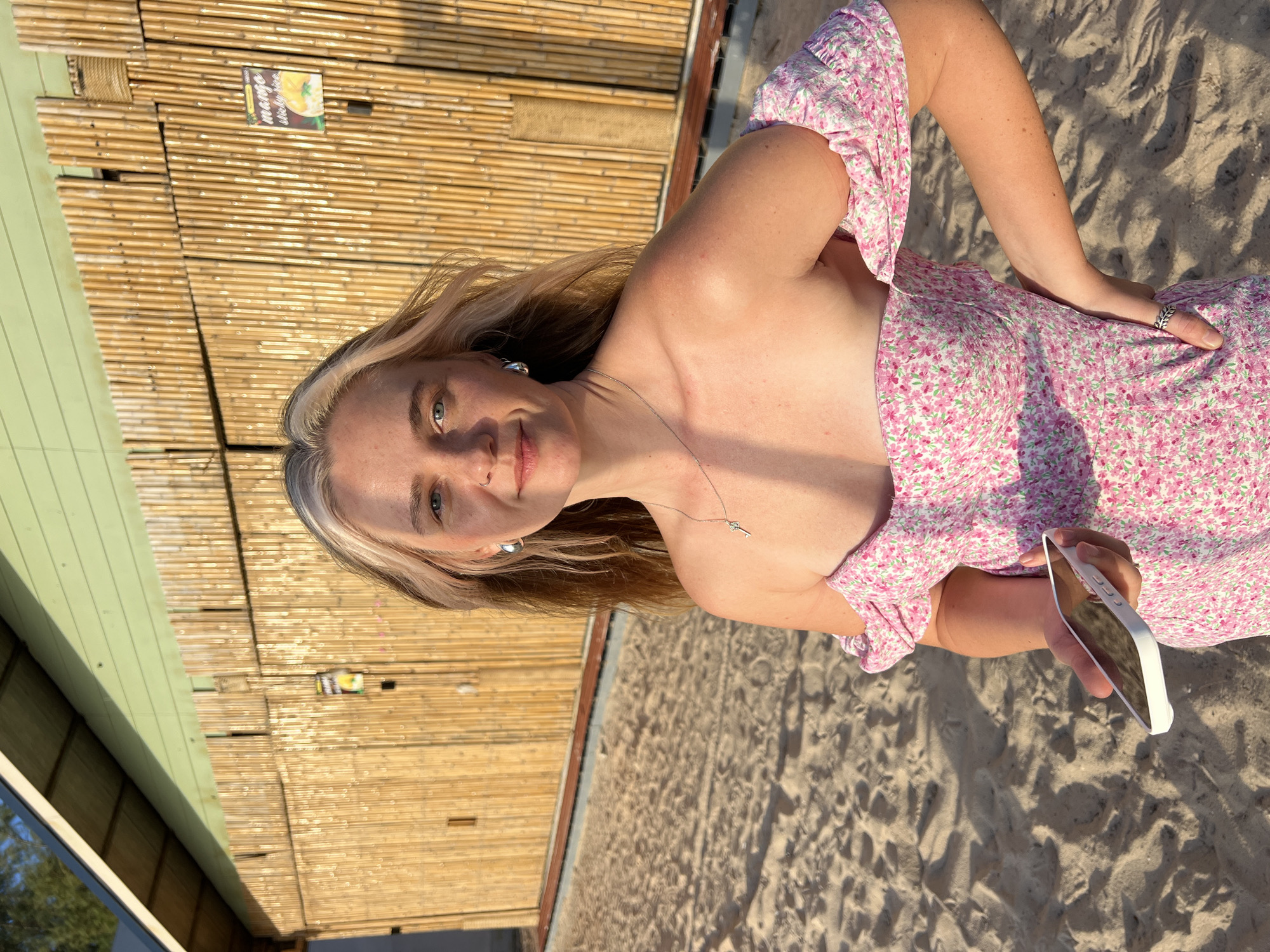 A young woman with blonde hair and blue eyes is lying on a sandy surface, possibly a beach. She is wearing a pink floral print dress and a silver necklace with a key pendant. She has a ring on her left hand. She is holding a white smartphone in her right hand. The background shows a wall made of bamboo and a green structure. There are shadows on the sand, suggesting it is daytime. The sunlight appears warm, hinting at either morning or late afternoon. The woman's expression is relaxed and her body language is casual. There is a small sign on the bamboo wall with some text that is partially visible. The overall scene conveys a sense of leisure and outdoor enjoyment.