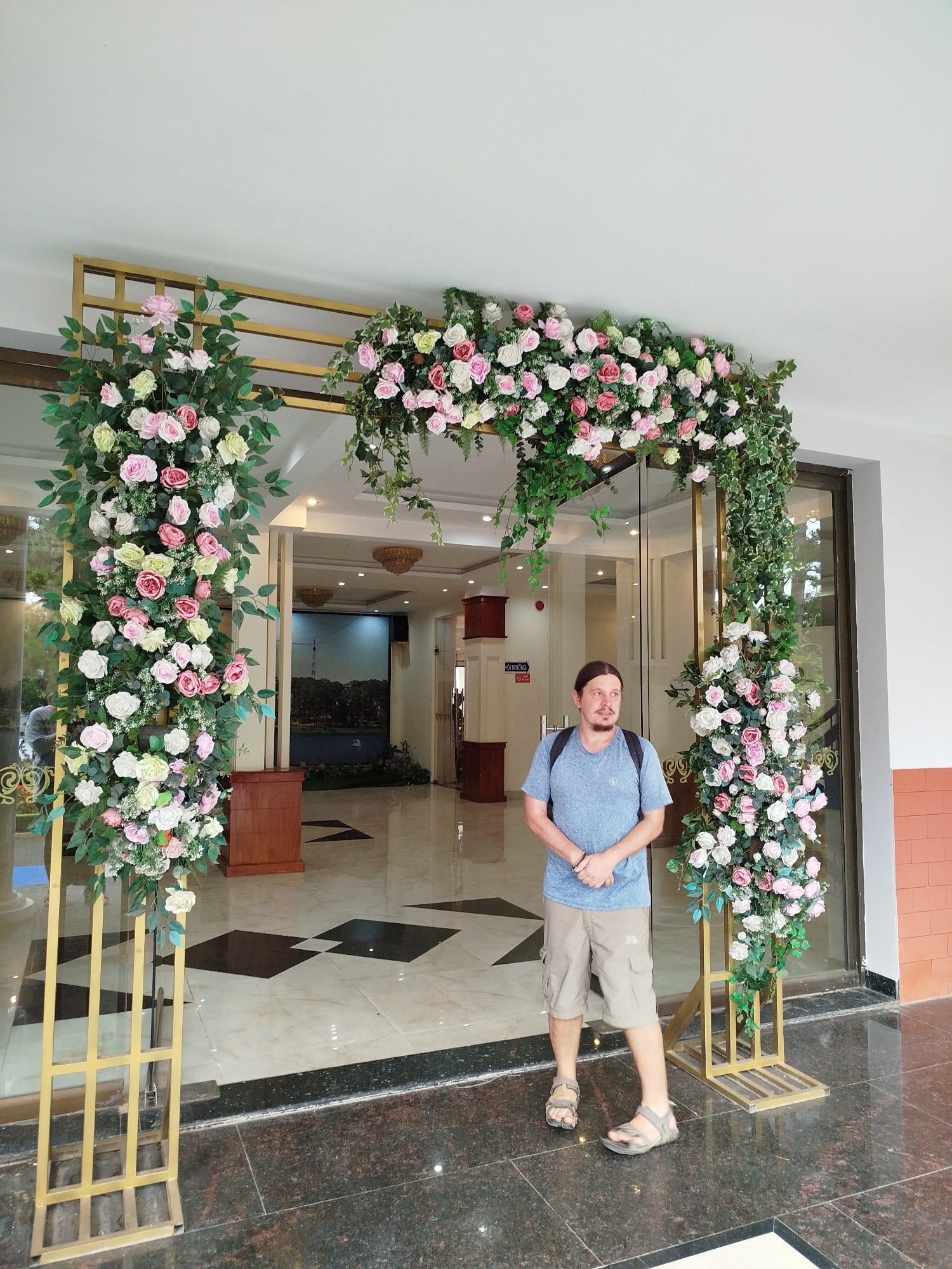 A man stands in front of a floral archway at the entrance of a building, possibly a hotel or restaurant, in Da Lat, Vietnam. The archway is decorated with artificial pink, white, and yellow roses and green foliage. The man is wearing a light blue t-shirt, khaki shorts, and sandals. He has a backpack on his shoulders. The background shows the interior of the building, with a large mural of a lake and trees on the wall. The flooring is a mixture of light-colored tiles with dark diamond patterns and dark polished stone. The lighting suggests it is daytime.