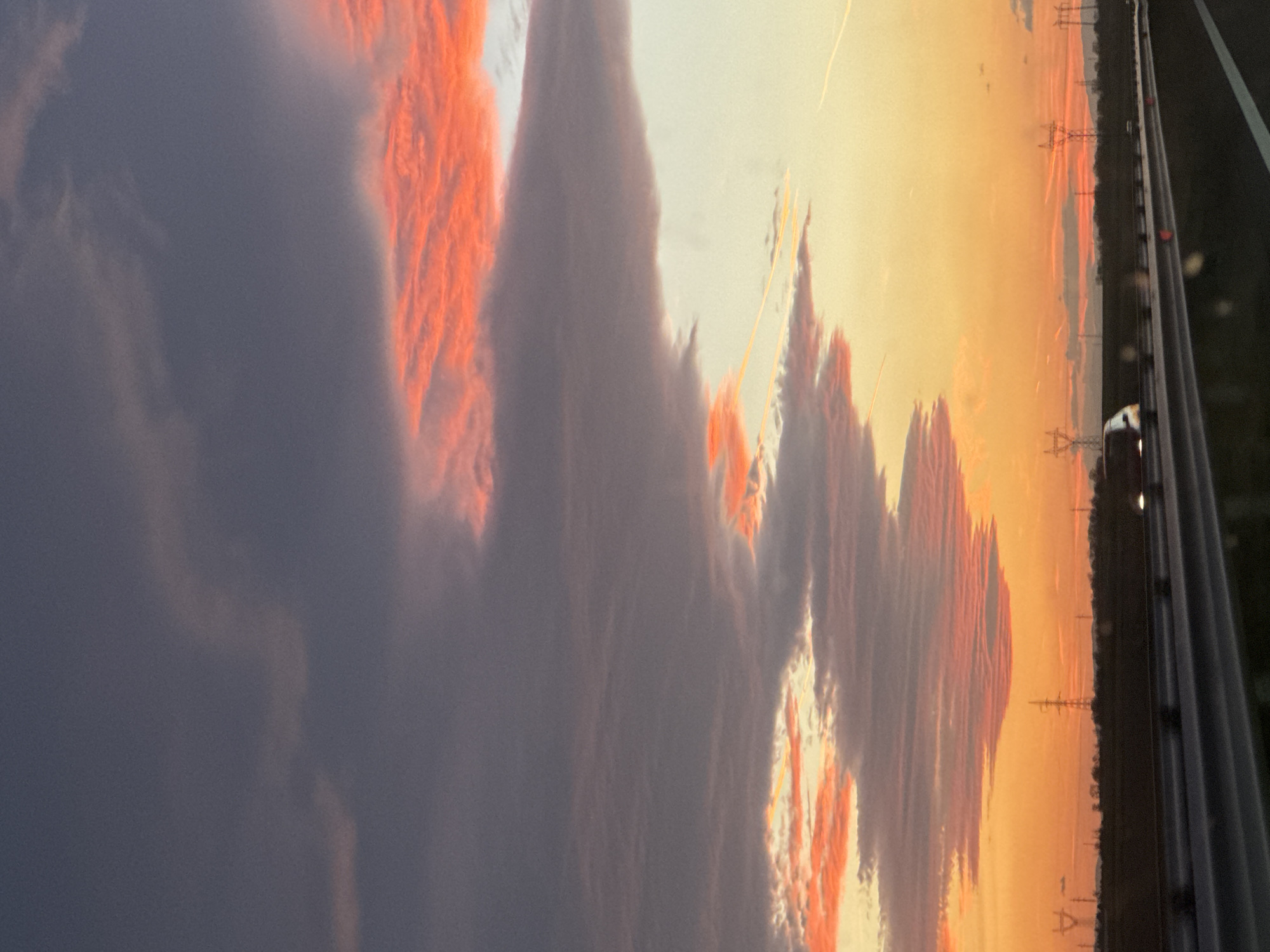 The image shows a sunset view captured from inside a moving vehicle, likely on a road near Malenovo, Bulgaria. The sky is predominantly orange and yellow, with streaks of orange-lit clouds against a gray cloud mass. The road infrastructure, including the guardrail and possibly a lane marker, is visible on the right side. Utility poles and overhead power lines are silhouetted against the sunset in the distance. A vehicle with headlights on is visible ahead on the road. The landscape includes trees. The overall scene suggests twilight or dusk.