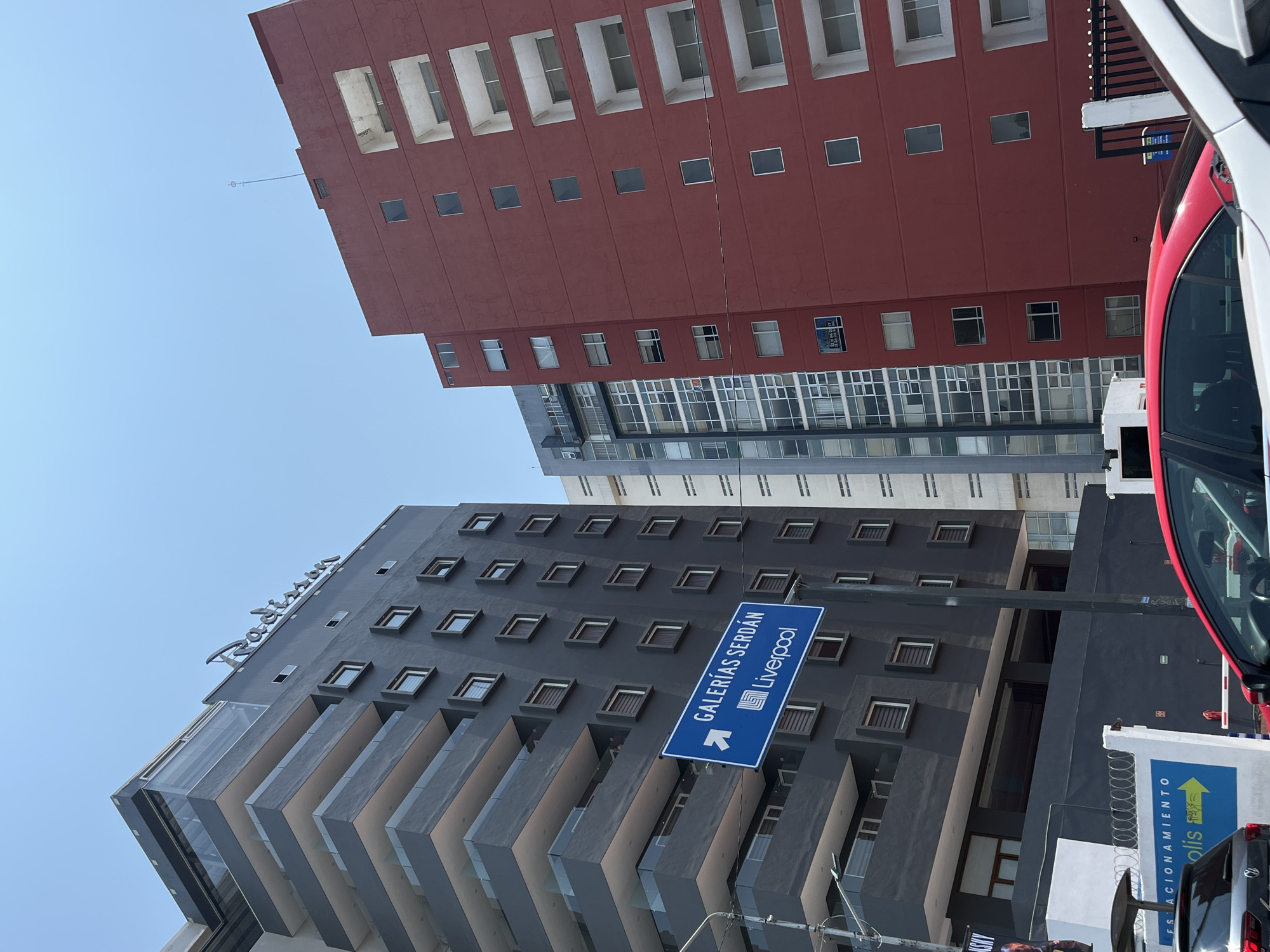The image shows a street scene in an urban environment. The sky is clear and blue. Several tall buildings are visible, varying in color. One building is reddish-brown with square windows, and another is gray, with a Radisson logo visible near the top. A blue street sign points towards "Galerías Serdán" and "Liverpool." There is a red vehicle on the right side of the frame. Also on the right is a partial view of a white fence with a yellow arrow.