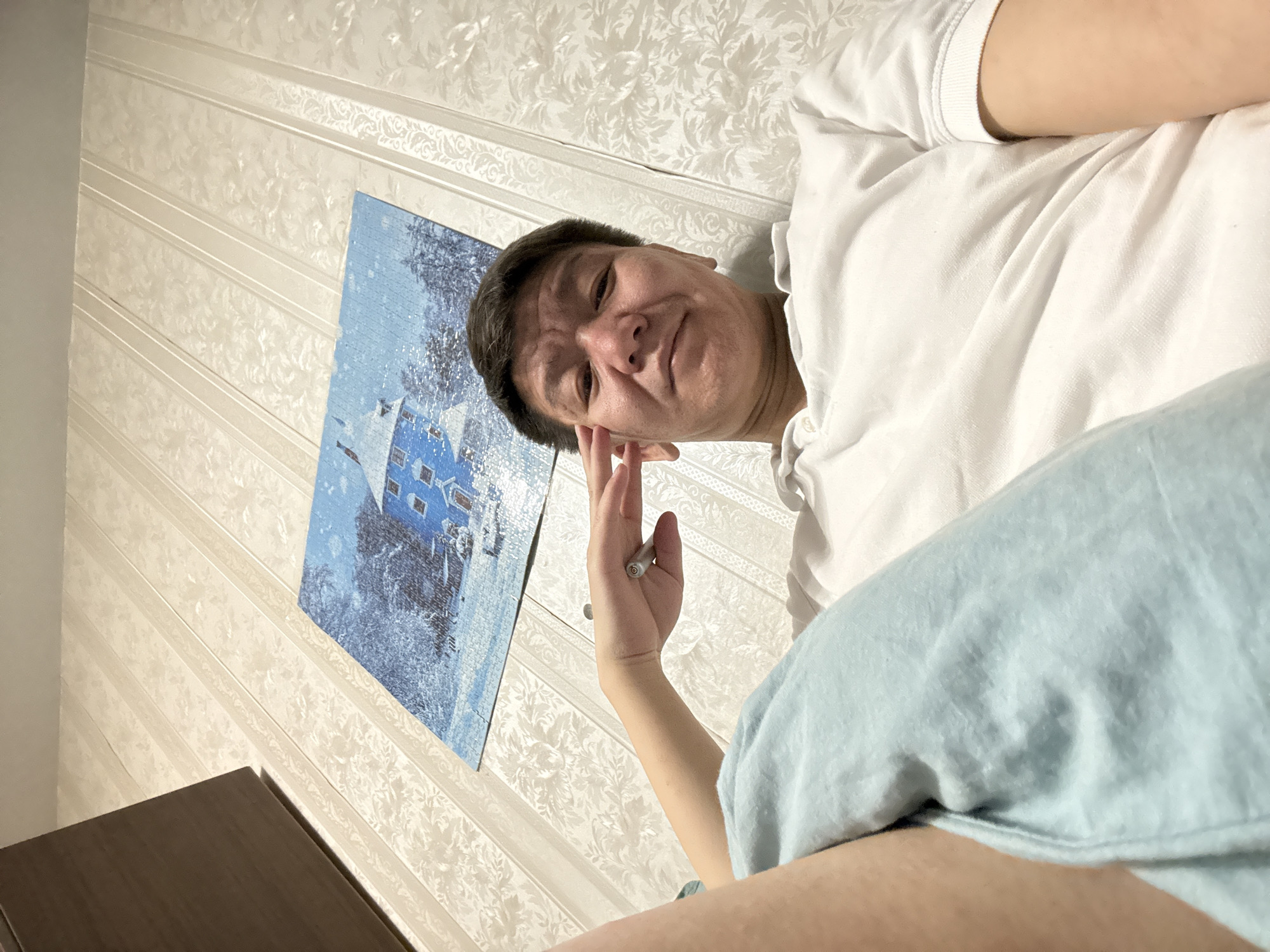 A middle-aged man is pictured from a low angle in an indoor setting. He is wearing a white polo shirt and has short, dark hair. His right hand is raised, with a pen visible between his fingers. A light blue blanket or sheet is draped over his lower body, and a portion of a dark brown piece of furniture is seen in the lower left corner.
Behind the man is a wall with patterned wallpaper in a light color. A framed picture is hung on the wall, showing a blue-toned image of a house or building in a snowy landscape.
There is no information in the image to determine that the image was taken in Khimki, Russia.