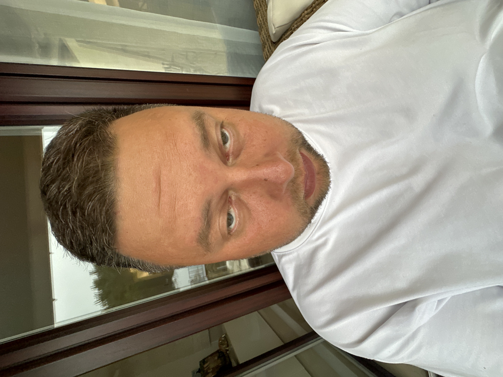 An adult man is featured in a close-up, chest-up shot, looking directly at the camera with a neutral expression. He has short, dark brown hair with visible grey strands, particularly around the temples, and light stubble on his chin and jawline. His eyes are brown and directly engaged with the viewer. He is wearing a plain white long-sleeved shirt, which appears to be made of a smooth fabric, showing various folds and creases.
The setting places him either indoors or on a covered patio or balcony, with a dark brown wooden window or door frame visible behind him. Through the glass pane, a diffused outdoor scene suggests daytime with overcast or partly cloudy weather. Green foliage, likely trees, can be vaguely discerned beyond the window, indicating an outdoor view. A light-colored, possibly woven, chair or cushion is partially visible on the right side of the frame. The overall context suggests a casual portrait taken in Sant Feliu de Guíxols, Spain. No visible text is present in the image.