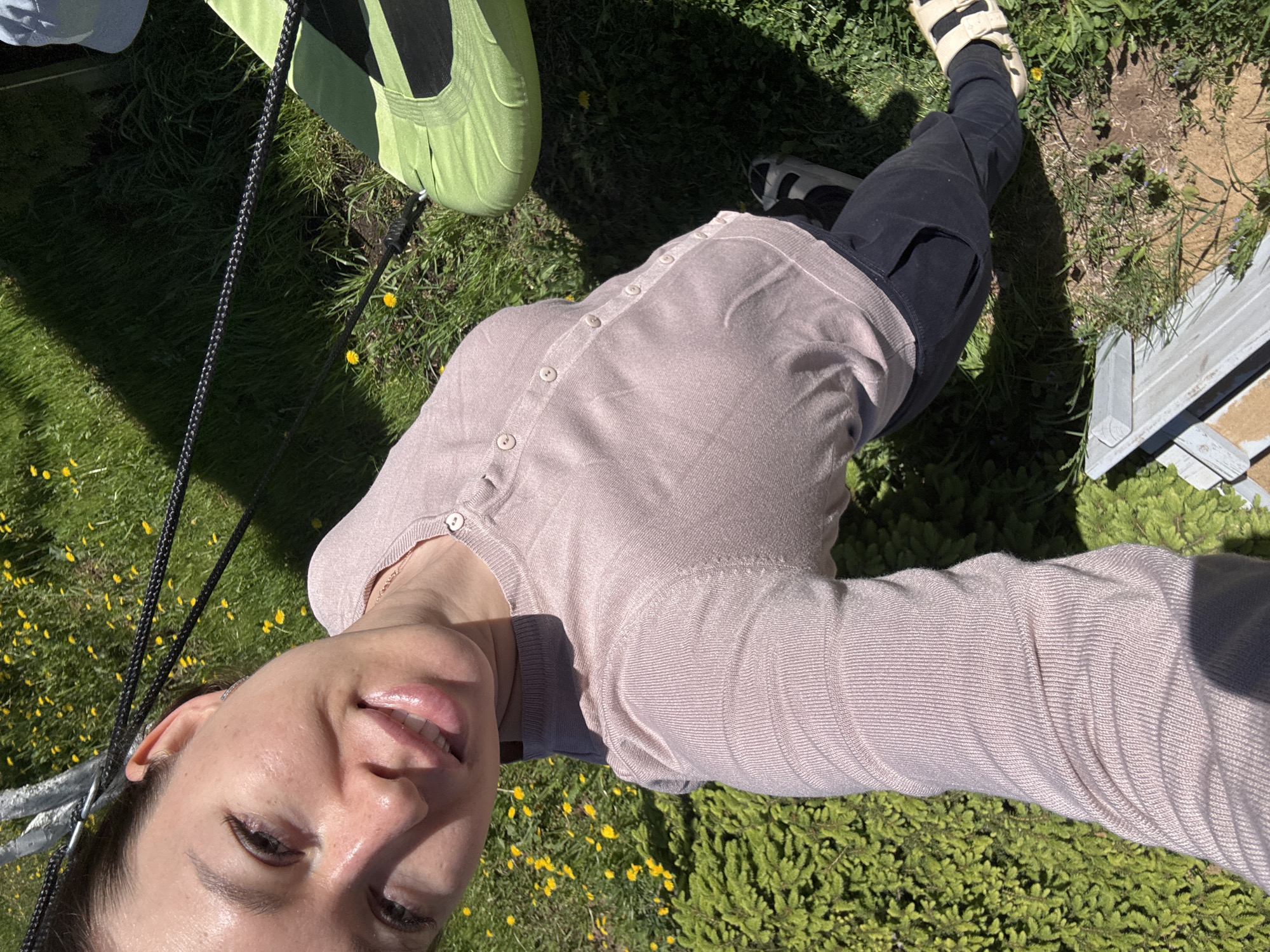 A woman takes a selfie outdoors. She is wearing a light pink button-down shirt and black cargo pants. Her footwear consists of one white sandal and one black and white athletic shoe. She is lying on a grassy surface with yellow flowers. A green hammock hangs nearby, supported by a black rope. A wooden structure is partially visible in the background. The location is likely a yard or park-like setting within Ферма, Russia, based on the user's query.