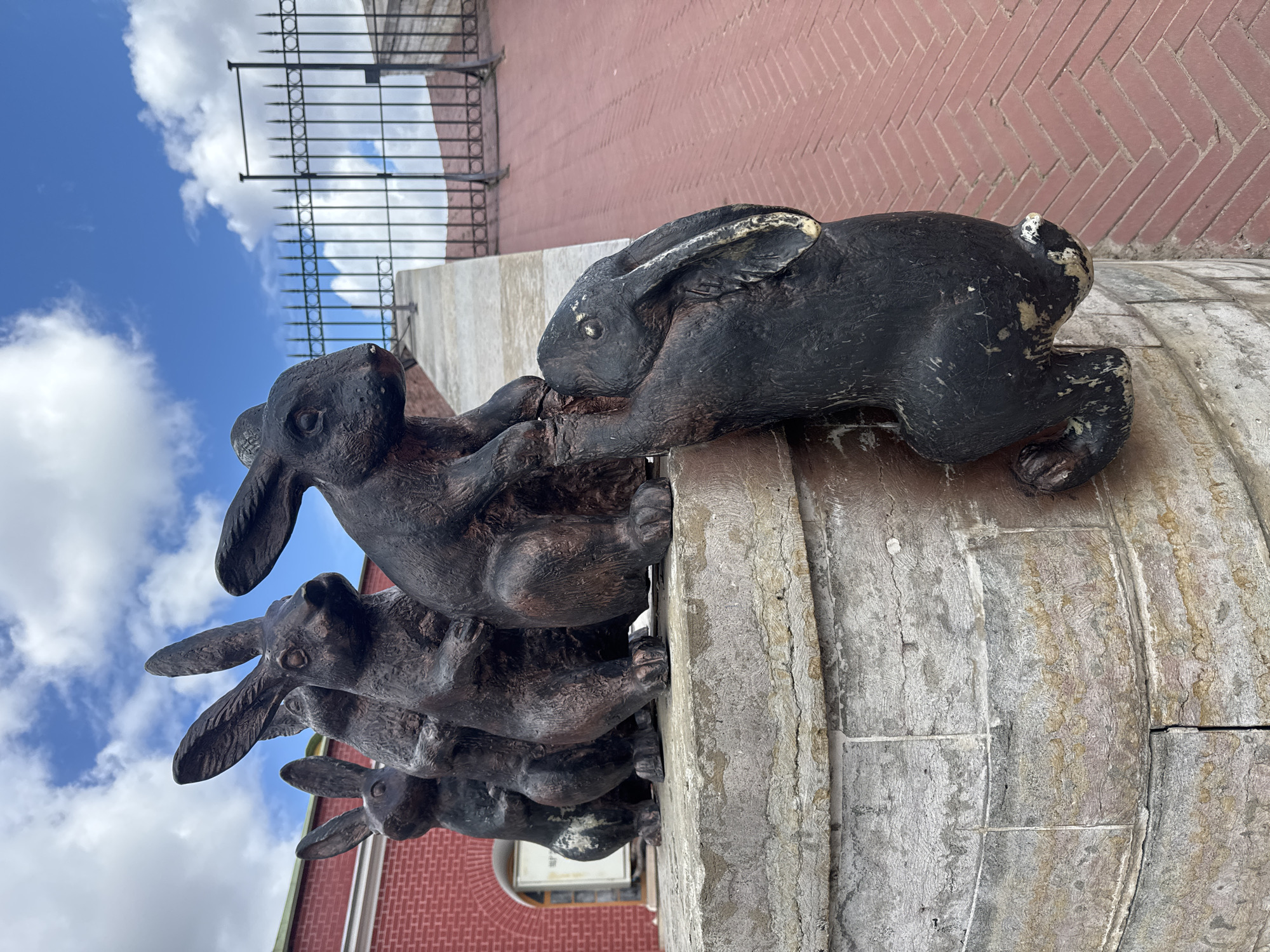 A low-angle, outdoor photograph features a sculpture of stacked rabbits in Saint Petersburg, Russia. The sculpture, made of a dark metal, depicts four rabbits, one on top of the other. The bottom three rabbits are positioned in a stack, while the top rabbit is lying horizontally across the top of the stack. The sculpture is placed on a weathered, cylindrical stone base.
Behind the sculpture, there's a brick building with a visible section of herringbone-patterned brickwork. A black metal fence is situated above the building. The sky, visible in the background, is blue with scattered white clouds.