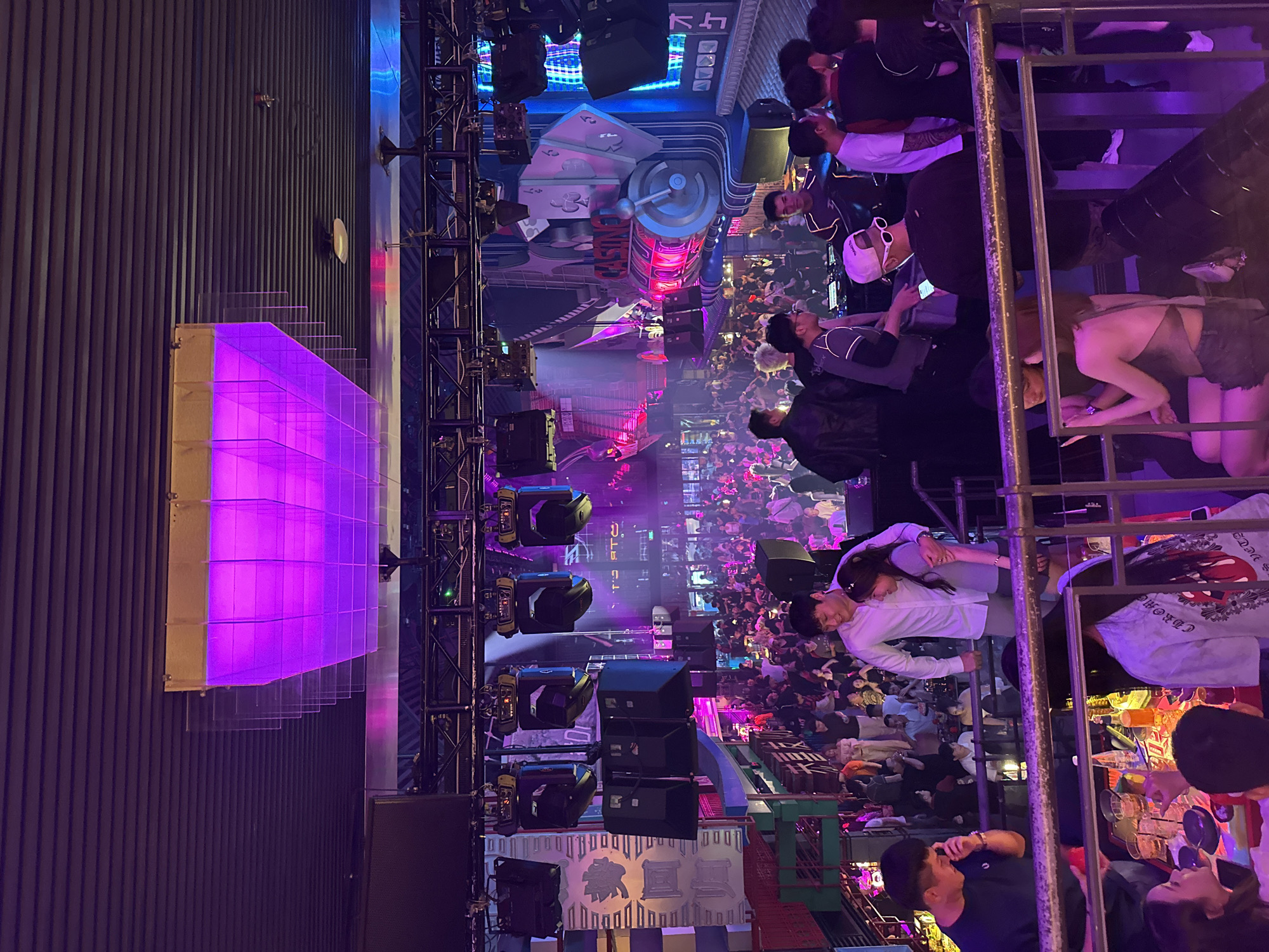 An indoor nightclub scene in Thủ Đức, Vietnam, is depicted from an elevated perspective. The venue is filled with a large crowd of people, many of whom are dancing or socializing. Above the crowd, elaborate lighting rigs with various spotlights and speakers are suspended from the ceiling. A prominent feature is a large, illuminated structure with pink and purple lighting, designed to resemble a stacked, grid-like panel. The walls are decorated with vibrant, cartoon-like graphics and architectural elements, including depictions of playing cards and a spinning slot machine, suggesting a playful, entertainment-themed atmosphere. In the foreground, a railing separates the observer's viewpoint from a group of people gathered around tables laden with drinks. One couple in the mid-foreground stands close together, with the man's arm around the woman's shoulders. The overall ambiance is lively and energetic, characterized by dynamic lighting and a dense gathering of patrons.