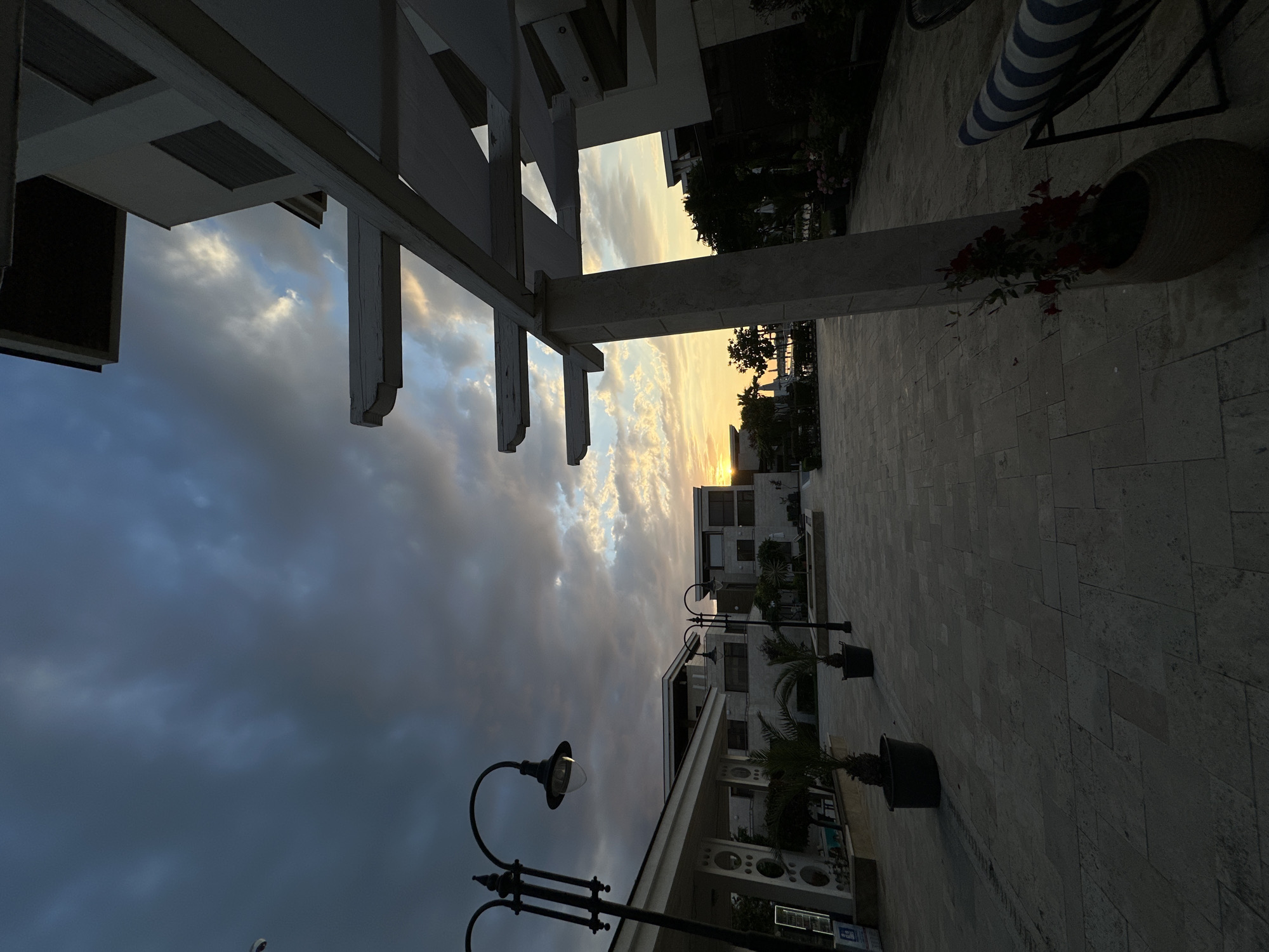 A paved courtyard area with modern architecture is captured during sunset in Sozopol, Bulgaria. The sky is dramatic with dark, heavy clouds interspersed with lighter patches where the sun is setting, casting a warm orange and yellow glow. In the foreground, a large, off-white tiled patio leads towards buildings. To the left, a white building with decorative circular elements is partially visible, along with a tall, black lamppost. Further into the courtyard, more contemporary buildings with dark-tinted windows are seen. Several potted palm trees are placed strategically around the courtyard. A white pergola structure, made of beams and fabric canopies, dominates the right side of the frame, casting shadows on the patio. In the far distance, trees and possibly water are visible. The scene is devoid of people, creating a quiet and somewhat somber atmosphere as day transitions into evening.