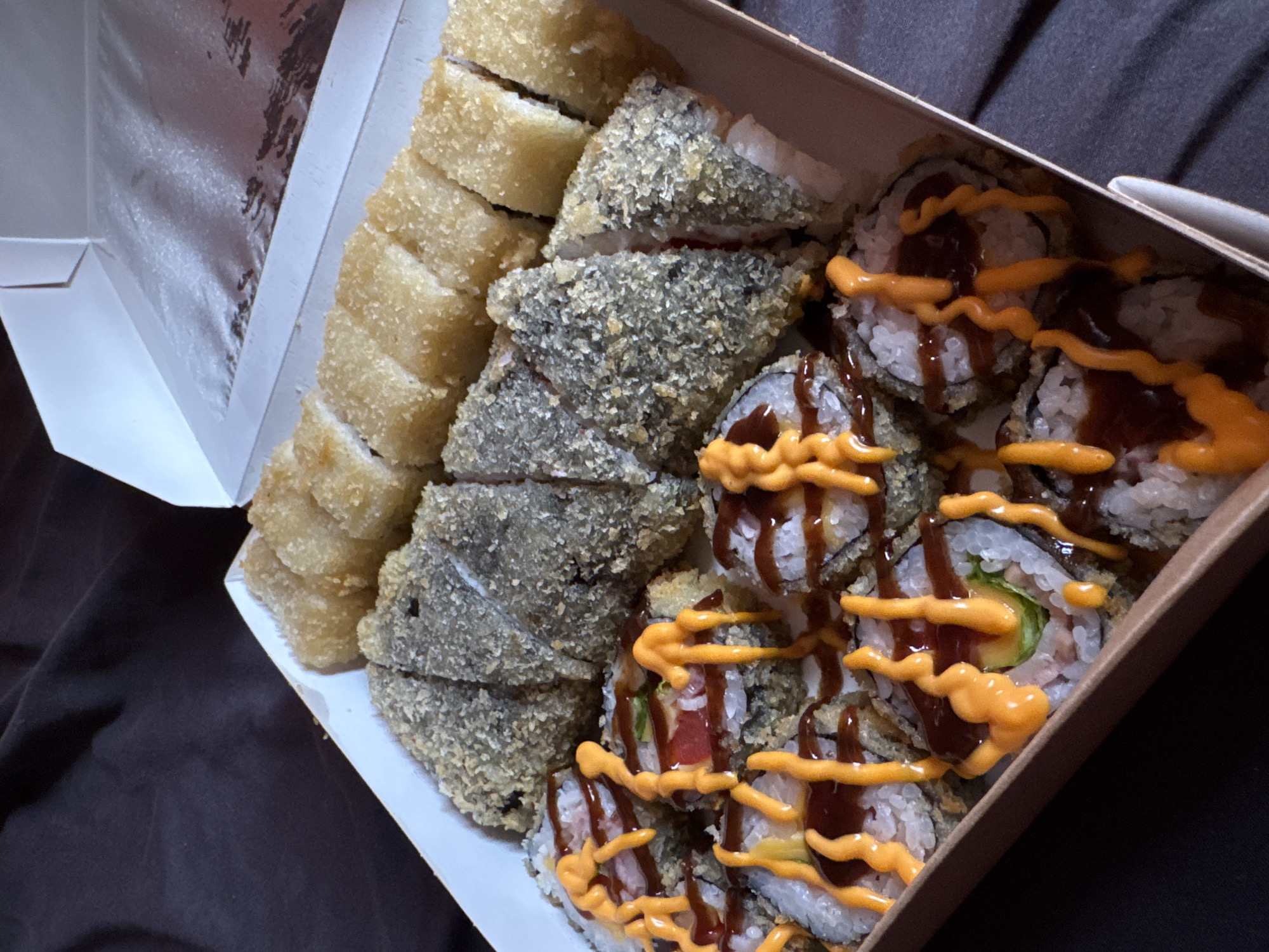 A partially opened white cardboard box containing sushi is positioned on a dark brown surface. The sushi is arranged in three rows. The first row features golden-brown, evenly cut sushi rolls. The second row consists of darker, rough-textured sushi rolls. The third row contains sushi topped with orange and brown sauces. The box is lined with a silver material. The surrounding environment is a dark brown surface. The location of the city of Chaikovsky, Russia, cannot be determined from the provided image.
