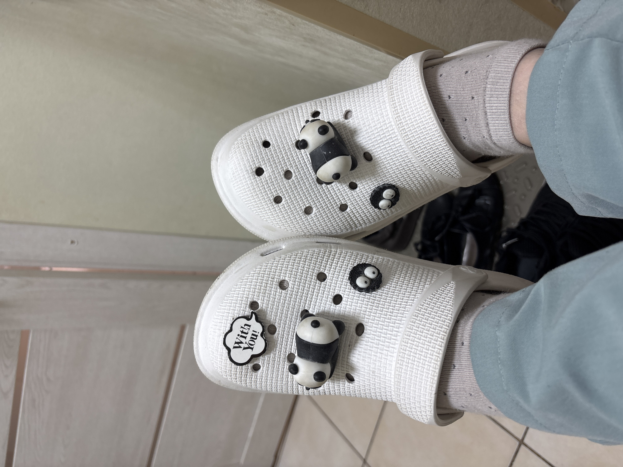 A pair of white Crocs-style shoes adorned with cartoon charms are visible from a top-down perspective. Each shoe has a panda charm and a two-eyed creature charm. One shoe also features a "With You!" charm. The shoes are being worn with light grey socks. The wearer also has on light blue pants. The setting appears to be an indoor location, with white walls and tiled flooring. In the background, black shoes can be partially seen. There is no information to determine the location as Vsevolozhsk, Russia.