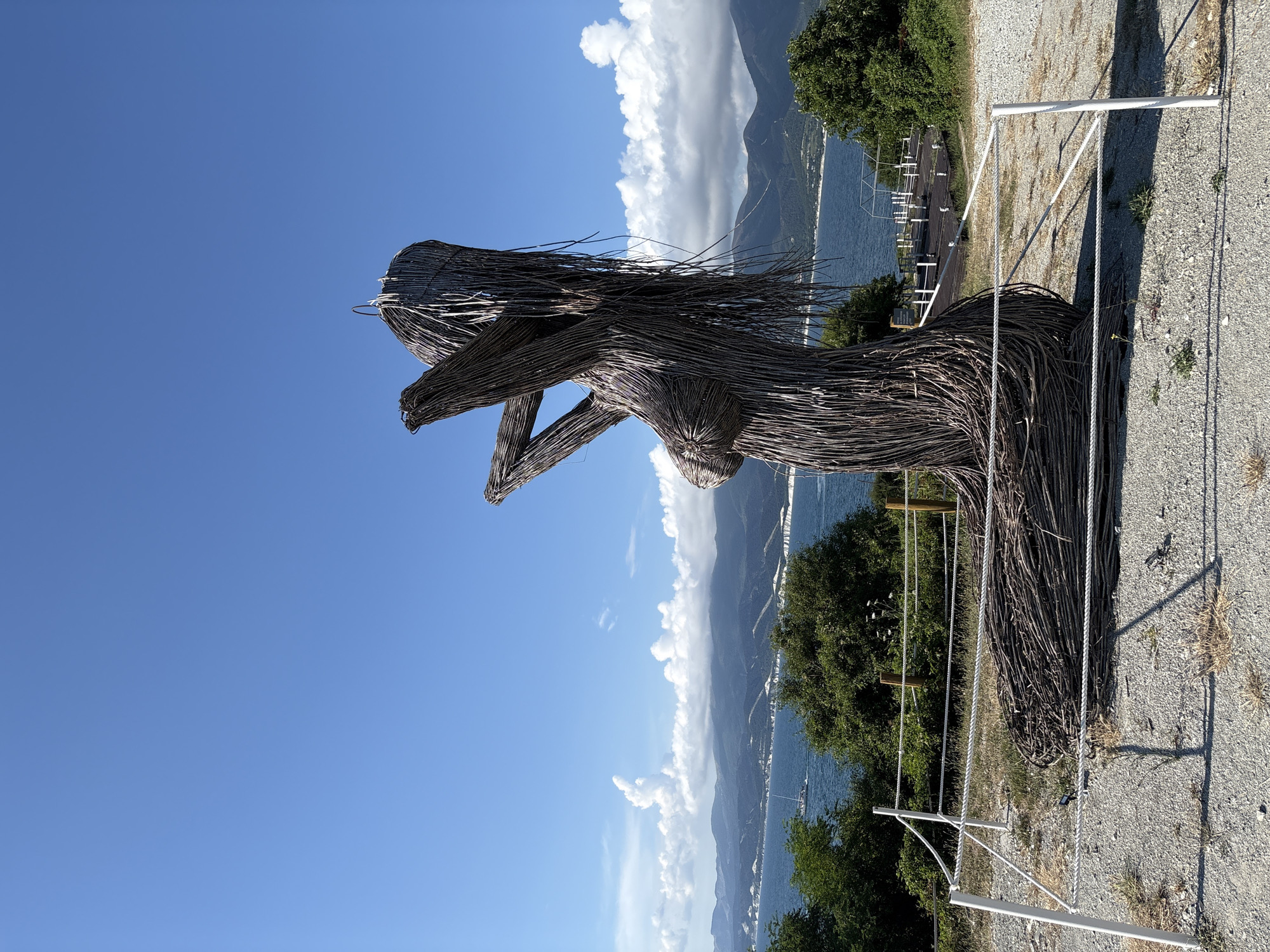 An outdoor sculpture, resembling a female figure made of interwoven branches, stands on a hillside overlooking the sea in Kabardinka, Russia. The sculpture is positioned in the foreground, with its right side facing the viewer. The figure has long, flowing "hair" and its arms are raised towards its head. In the background, a calm blue sea stretches towards a mountainous coastline under a clear blue sky dotted with fluffy white clouds. A white boat is visible on the water in the middle distance. Lush green trees and bushes surround the sculpture and dot the landscape. A white rope barrier with white posts encloses the sculpture, indicating it is an exhibit or point of interest. The ground is covered in gravel.