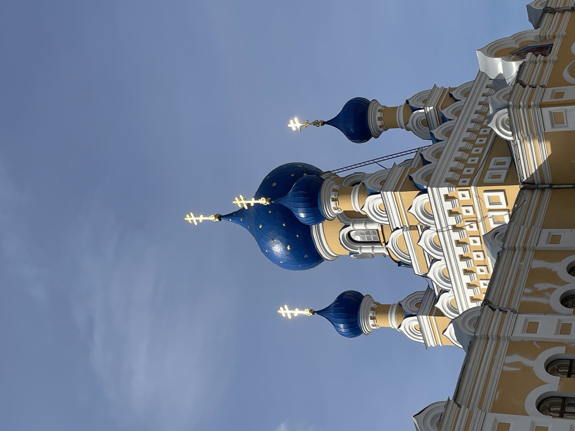 The image shows the upper portion of a yellow and white Orthodox church with multiple blue domes topped with golden crosses. The architecture is ornate, with decorative white trim and arched windows visible on the lower sections of the building. The sky is a clear, pale blue with a few wispy clouds. The lighting suggests it is daytime, with the sun reflecting off the golden crosses and the blue domes. A ladder can be seen leaning against one of the domes, indicating potential maintenance or cleaning work. The church's facade exhibits some signs of wear, with peeling paint visible in some areas. This church is identified as St. Nicholas Orthodox Church, located in Brest, Belarus. No people are visible in the image.