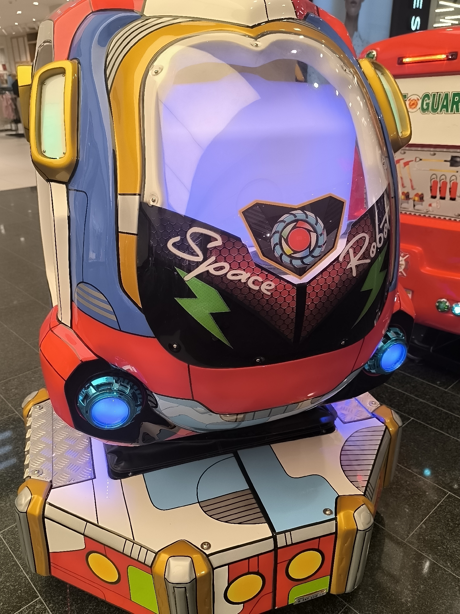The image shows a close-up view of a colorful, child-friendly amusement ride designed to resemble a robot, likely located in a shopping mall or entertainment center. The ride features a transparent bubble top with blue interior lighting, and its exterior is a vibrant mix of blue, red, yellow, and white with graphic designs including lightning bolts and the text "Space Robot." Two prominent blue LED lights are visible on the lower front of the ride. The base of the ride is also elaborately decorated with geometric patterns and colors. In the background, a red amusement ride with the word "GUARD" visible on it can be seen. The flooring appears to be dark, patterned tiles. The specific location of Klaipėda, Lithuania, is not discernible from the image content itself.