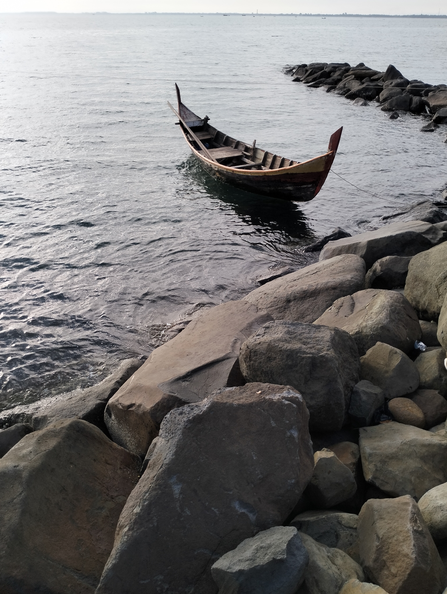 A wooden boat rests in calm water near a rocky shoreline. The location appears to be in Lhokseumawe, Indonesia. The boat is likely secured to the rocks. The water is slightly rippled with reflections of sunlight. Beyond the boat and rocks, the sea stretches towards the horizon under a bright, overcast sky. The rocks are large and unevenly piled.