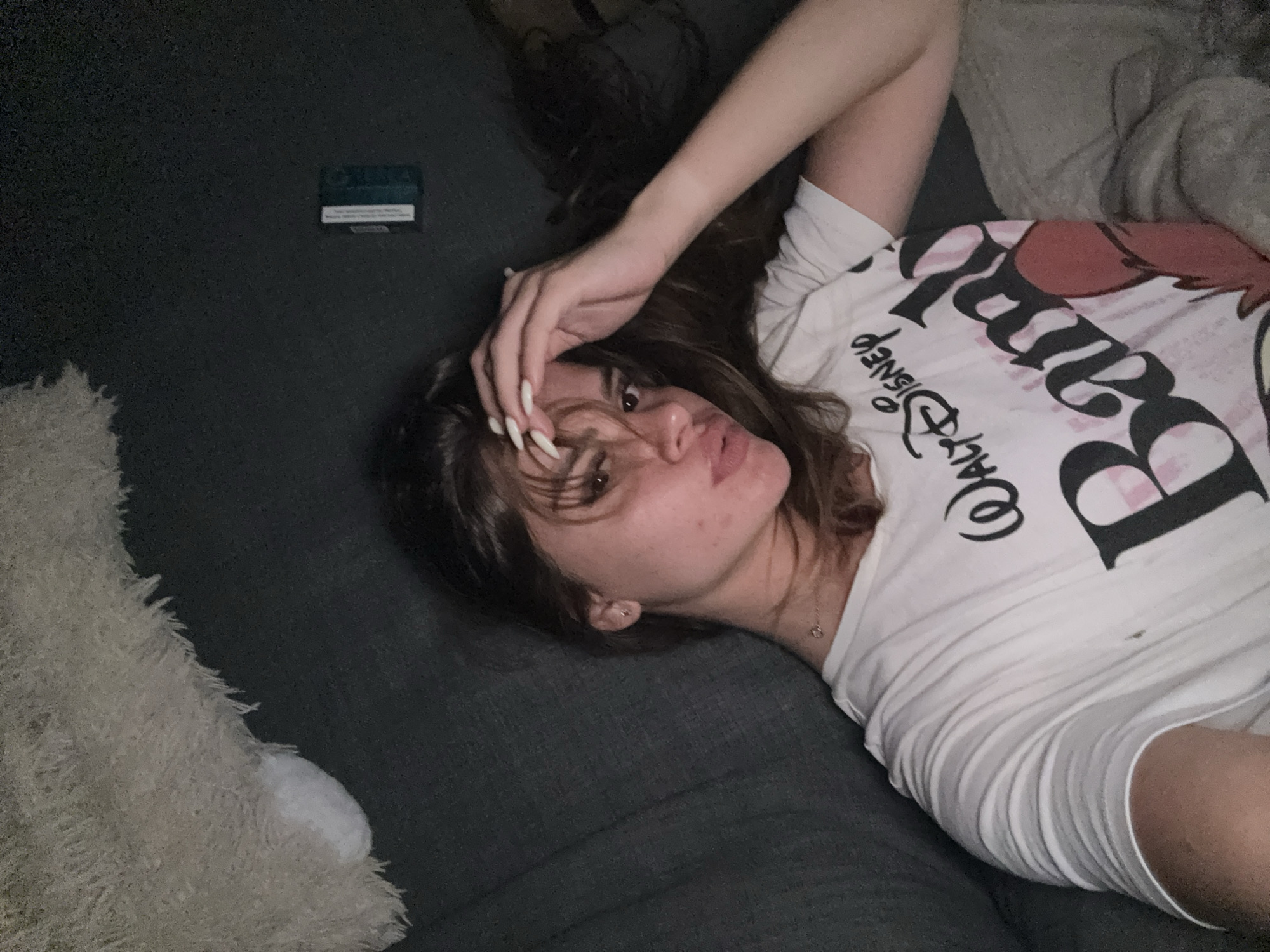 A young woman with brown hair and fair skin, wearing a white t-shirt with a "Walt Disney Bambi" graphic, lies on a dark-colored couch. She is looking towards the camera with her hand on her forehead. The environment appears to be indoors, with a furry throw blanket visible in the upper left corner. A small, rectangular object, possibly a pack of gum or a small box, is on the couch behind her to the right. The lighting suggests an evening or dimly lit setting. There is no indication of the location being in Plovdiv, Bulgaria.