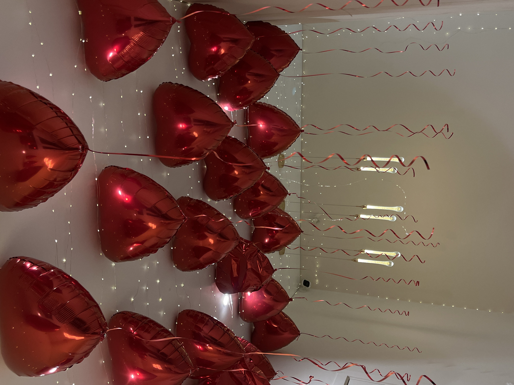The image shows a room decorated with red, heart-shaped balloons attached to the ceiling with red, curling ribbon. String lights also adorn the ceiling. No people are visible. The location is inferred to be in Neftekamsk, Russia, but no evidence in the image directly supports this location.
