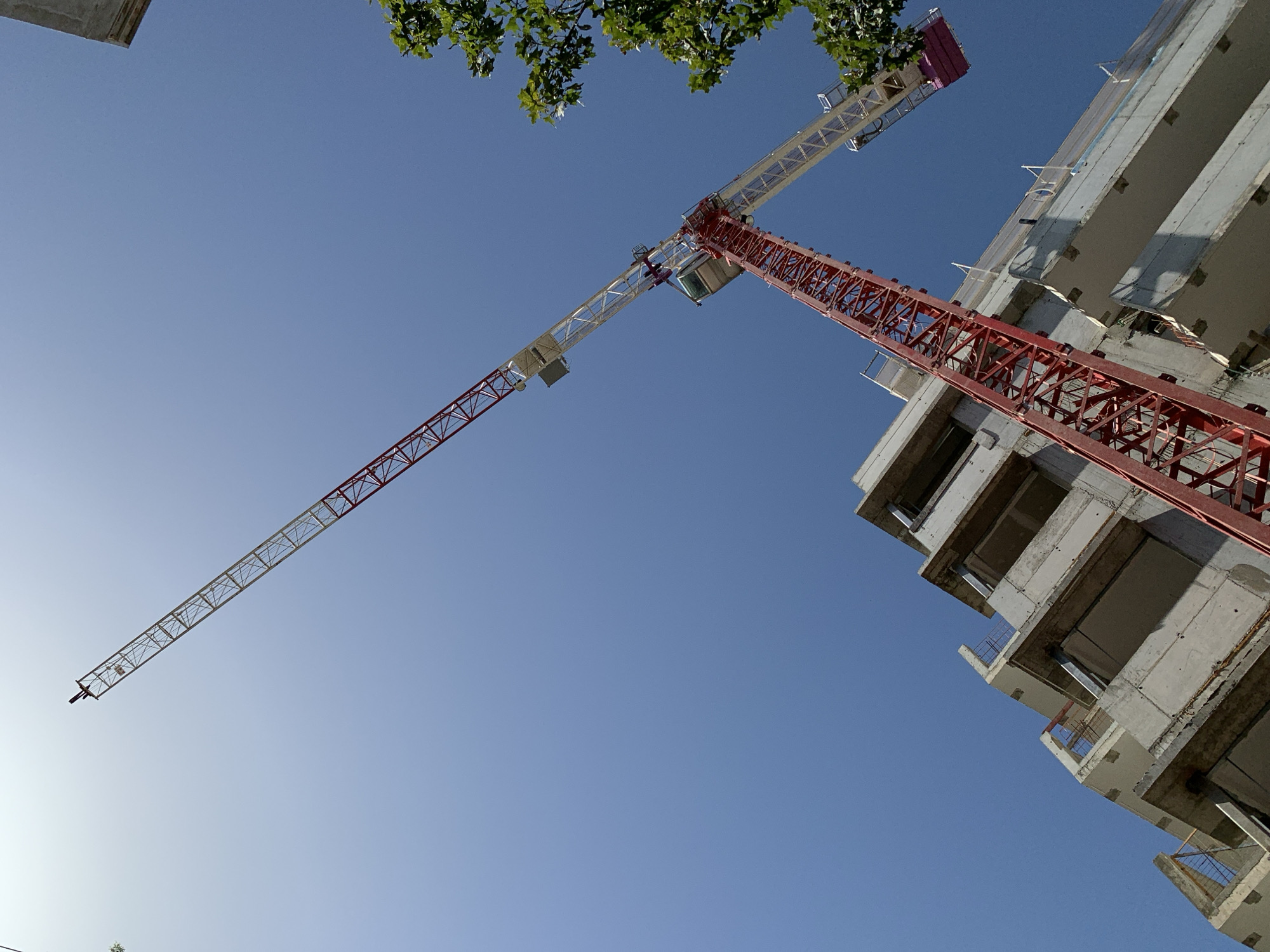 The image depicts a construction site in Tel-Aviv, Israel. A tall, red and white construction crane dominates the view, contrasting against the clear blue sky. The crane is positioned near a partially constructed concrete building, several stories high. Green foliage is visible at the top of the frame, suggesting nearby trees. The scene is taken from a low angle, looking upwards at the crane and building.