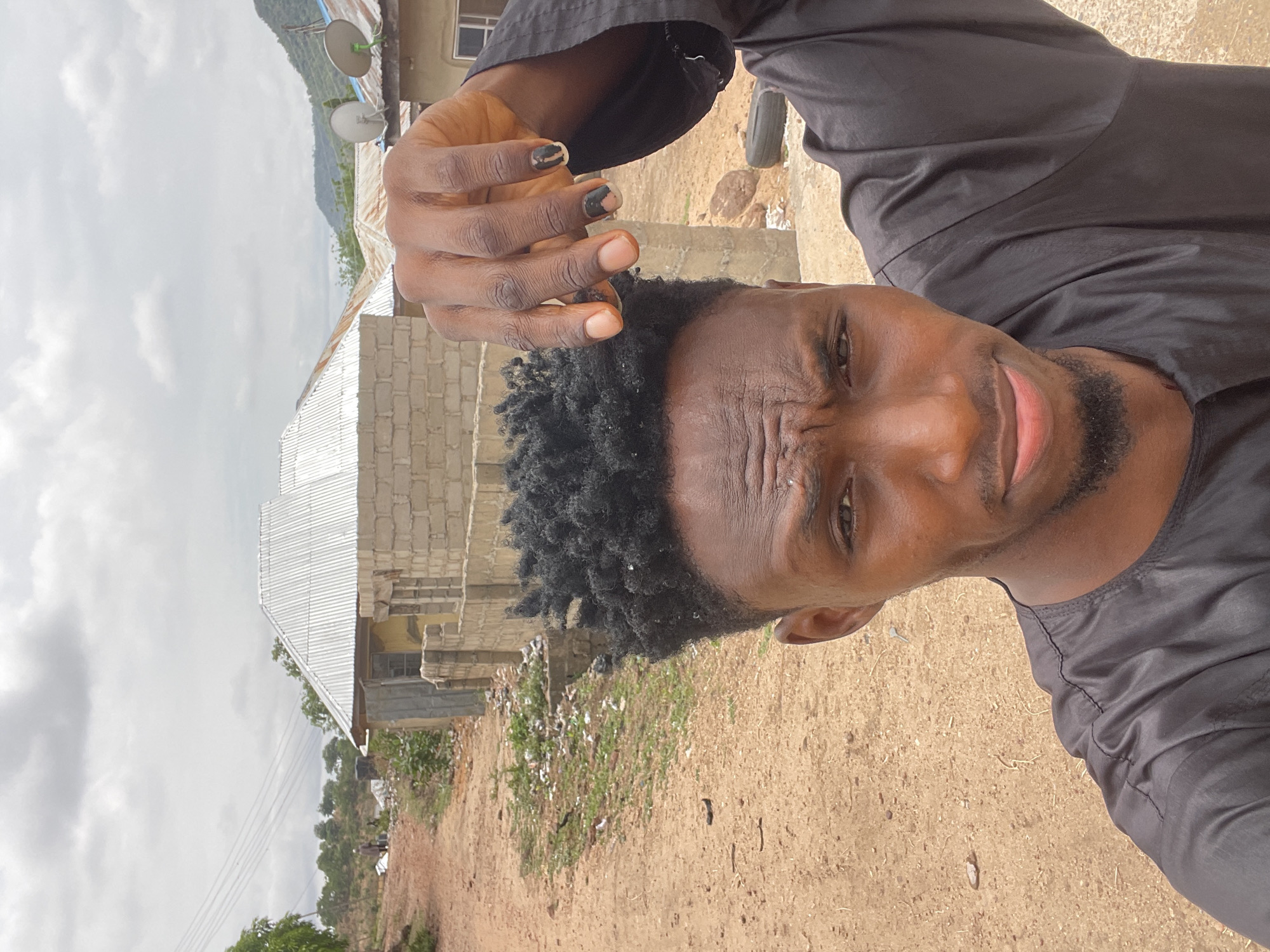 A man is posing for a selfie in Lokoja, Nigeria. He has dark skin, short dark curly hair, and a trimmed beard. He is wearing a black shirt. His left hand is raised near his head, with painted nails. Behind him is a partially constructed building with a corrugated metal roof. The surrounding area appears to be a dirt yard with sparse vegetation and some trash. In the background, there are more buildings, trees, and a hill or small mountain. The sky is overcast. Satellite dishes are visible on one of the buildings. Power lines are strung above the scene.