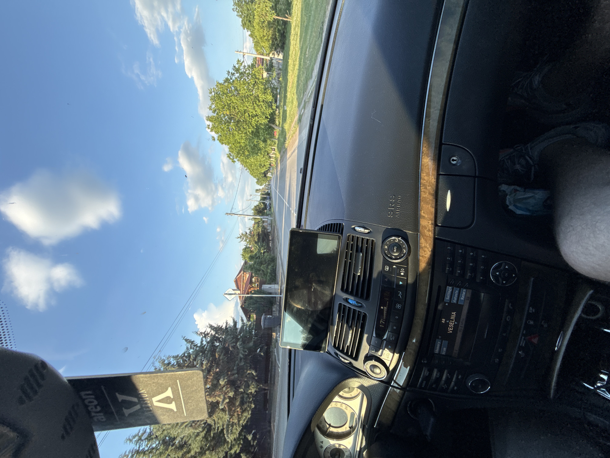 The image is taken from inside a vehicle, likely the driver's seat, looking out the windshield. The vehicle is moving on a street lined with trees and houses. The dashboard is visible, displaying the car's controls and information. The sky is blue with scattered clouds. The text on the dashboard indicates the name of the radio station is "VESELINA," and "SRS AIRBAG" is printed. The location is identified as Sarnevo, Bulgaria, based on the radio station name. An air freshener labeled "Areon" with a "V" symbol is attached to the windshield. A person's arm is visible on the right side of the image.