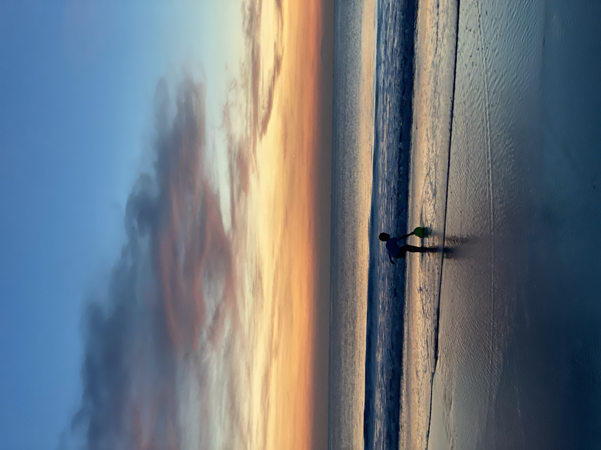 A person, likely a child, is standing on the beach with a green bucket in hand. The ocean meets the beach, with a small wave visible. The sky transitions from blue to orange/yellow, suggesting a sunset. The photo was taken at the beach in Kuta Selatan, Indonesia.