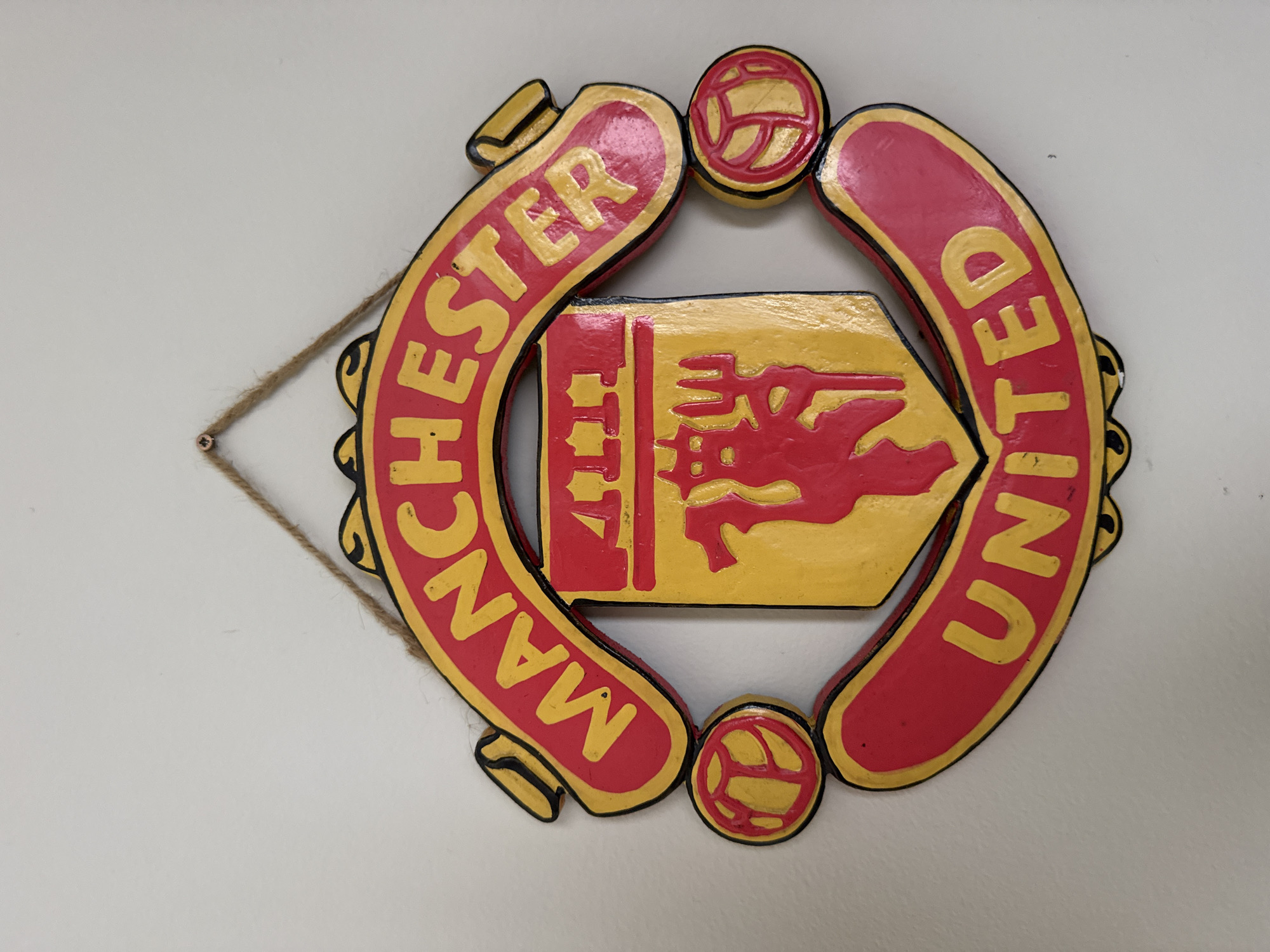 A Manchester United logo plaque hangs on a white wall. The plaque features the words "MANCHESTER UNITED" in yellow letters on a red, horseshoe-shaped border with black trim. A yellow shield with a red devil emblem sits in the center. The plaque is hung with a piece of twine attached to the wall. The location in the city of Unknown, Unknown is not determinable from the image.