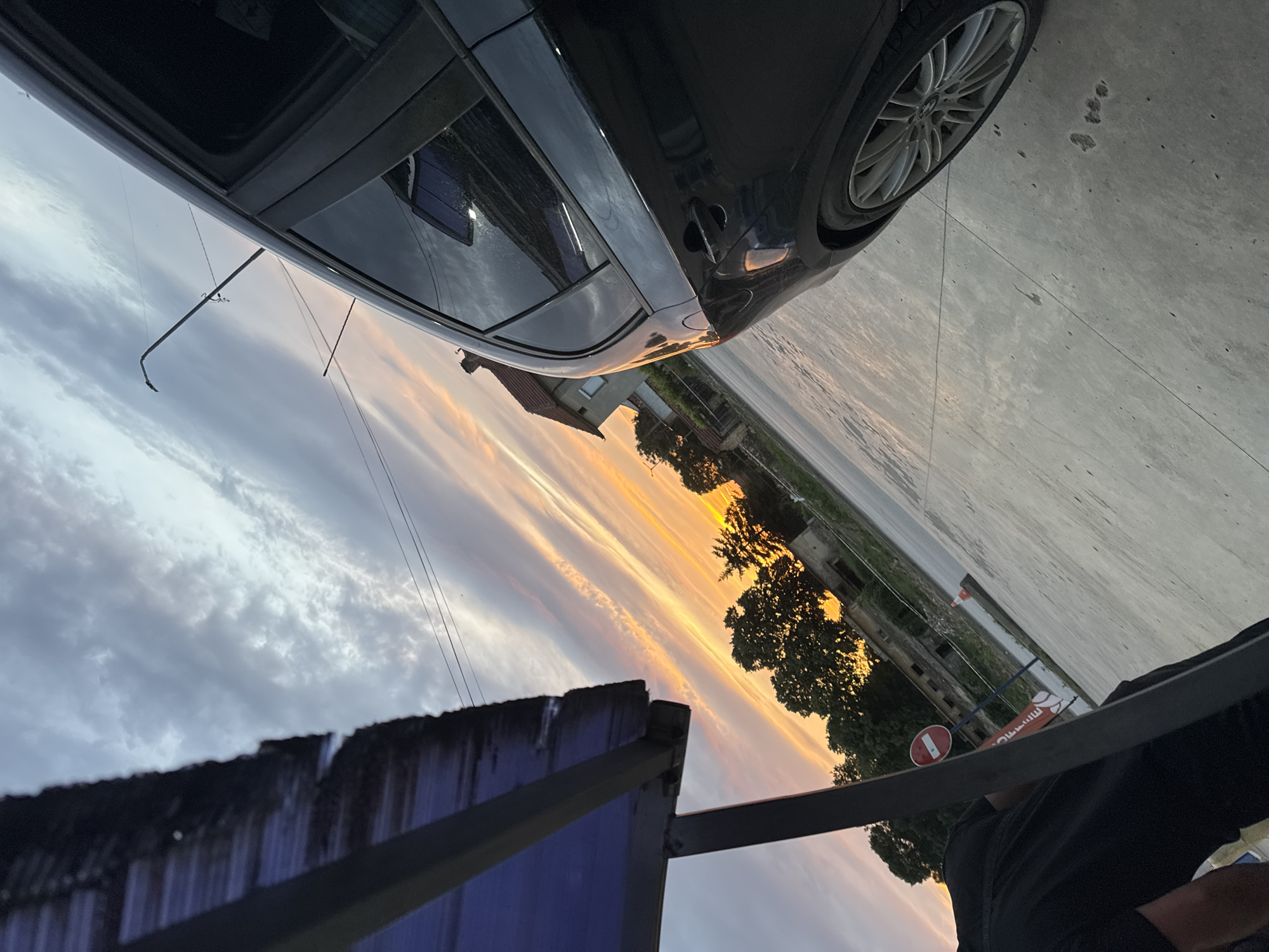 The image shows a sunset scene in Dolna Oryahovitsa, Bulgaria. A black car is parked on a concrete surface. In the background, there's a building and trees, with a colorful sunset sky above. A blue metal structure with dark trim is visible in the foreground, along with a person partially visible in the lower right corner wearing a black shirt. Also visible is a "no entry" traffic sign. The scene appears to be taken from a low angle.