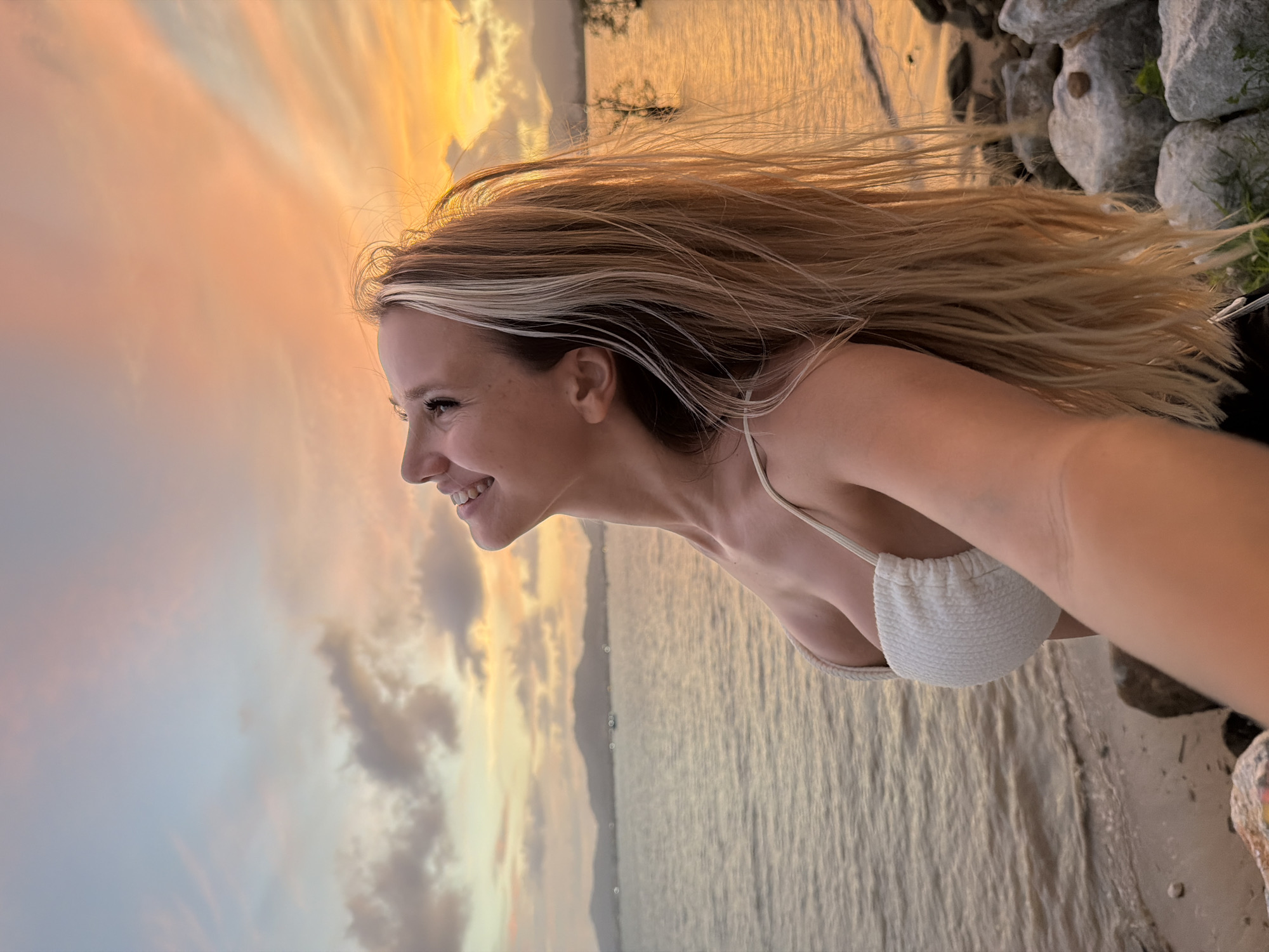 A woman with long blonde hair is standing on a rocky shore by the water at sunset. She is wearing a white bikini top and looking up at the sky with a smile. The sky is filled with colorful clouds in shades of orange, pink, and blue. The water is calm and reflects the colors of the sunset. The location is Wichit, Thailand.