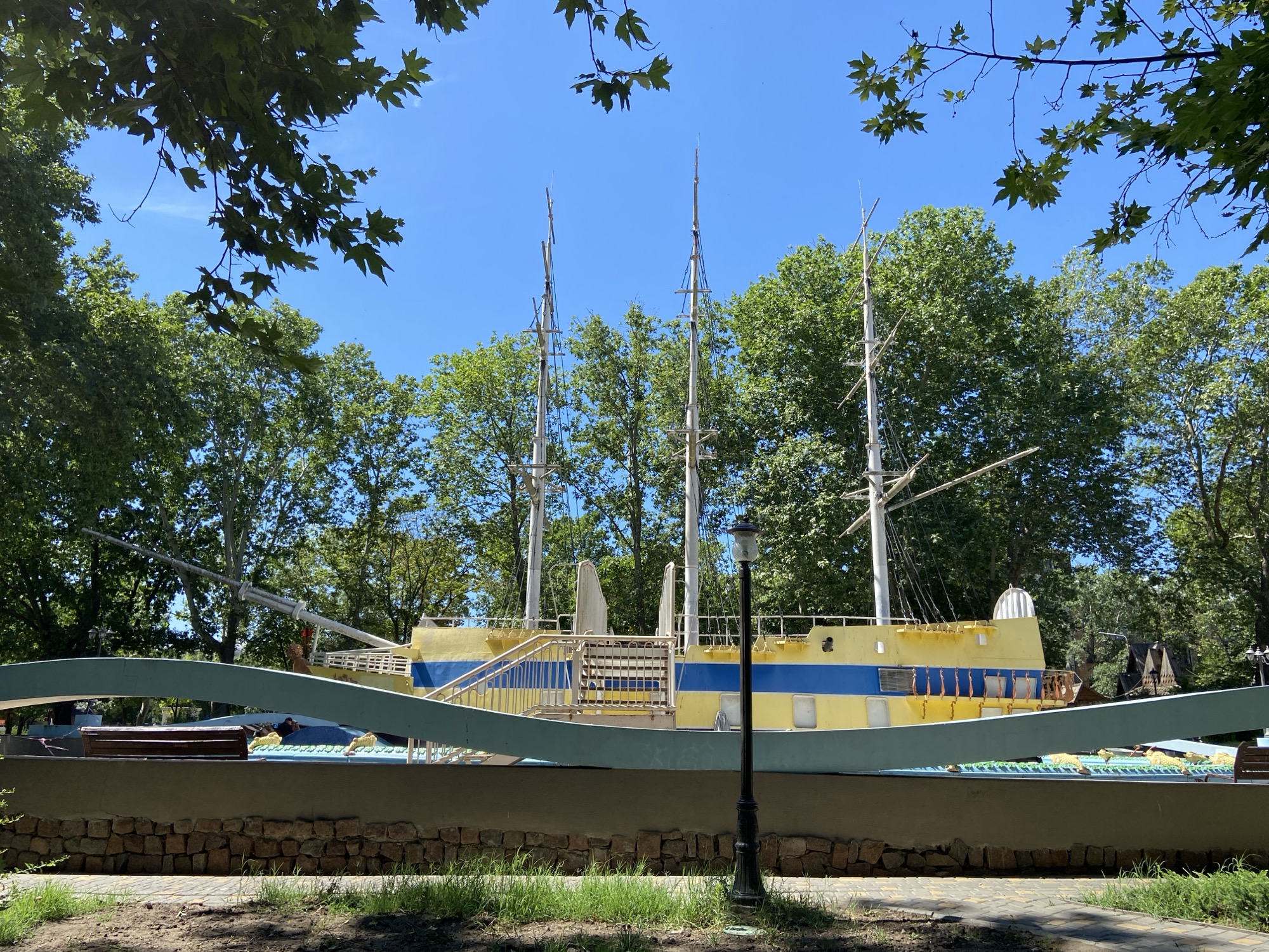 In an outdoor scene, a yellow and blue ship structure, likely a playground or attraction, is displayed against a bright blue sky. The structure features three masts, suggesting a sailing vessel. Lush green trees surround the ship, and branches with green leaves frame the top of the image. In the foreground, a stone wall with a paved pathway and grass is visible. A black lamppost stands to the right of the ship. The ship itself is decorated with blue stripes and has a light blue hull-like element in front of it. There are stairs leading up to the ship, and what appears to be a bench within the ship's structure. The overall scene suggests a park or recreational area.
