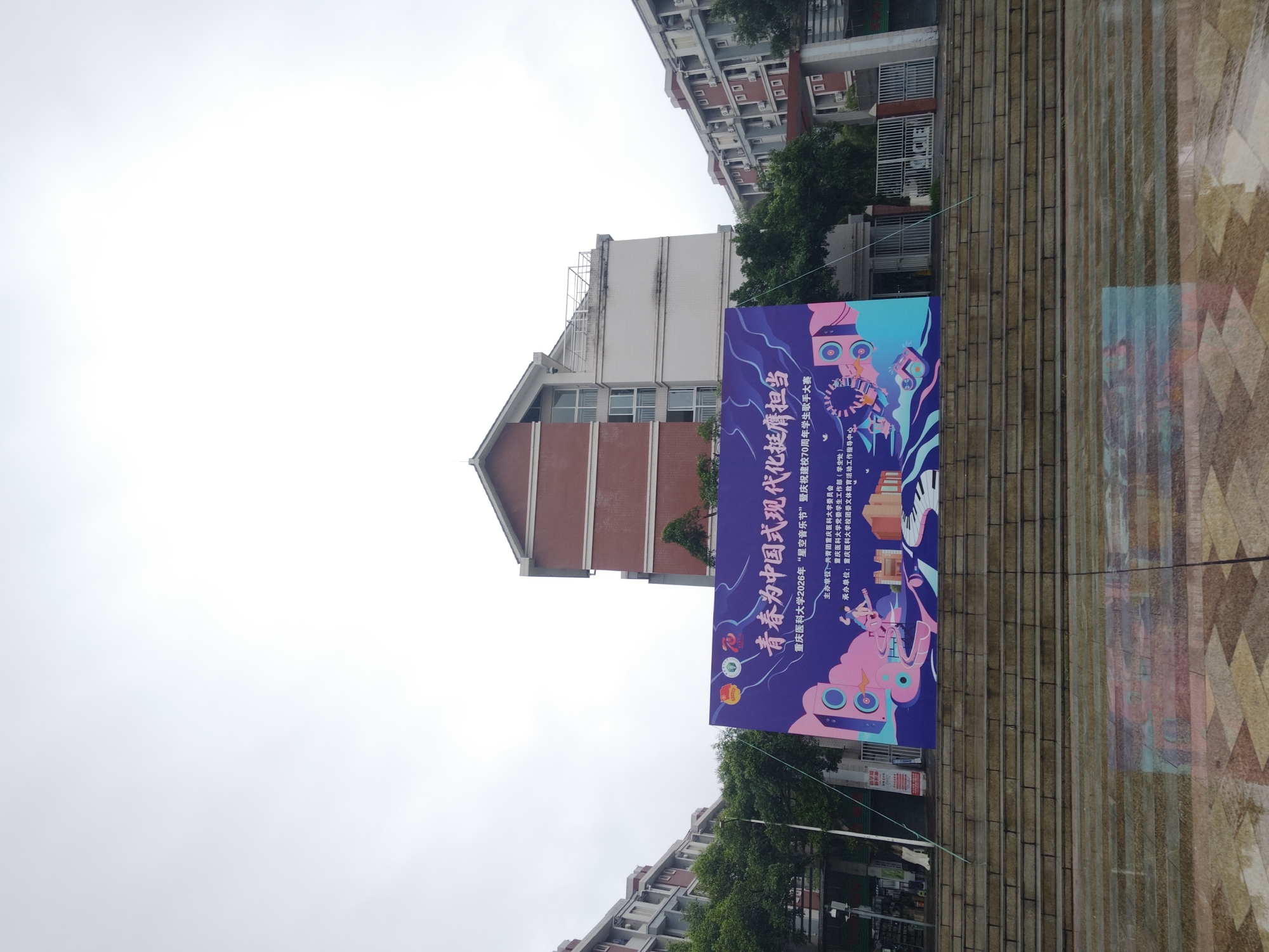 The image depicts an outdoor scene at Chongqing Medical University, situated within the Chongqing High-tech Zone, China, under an overcast sky. A large, horizontally oriented banner, predominantly purple and blue with stylized, vibrant graphics illustrating music festival elements such as speakers, a guitarist, and a piano keyboard, is centrally positioned. The banner's text announces the "Chongqing Medical University 2026 'Starry Sky Music Festival' Celebrating the 70th Anniversary of the School's Founding Student Singer Competition," organized by the Communist Youth League Chongqing Medical University Committee and the University Student Affairs Department.
The banner is suspended above a wide, multi-tiered concrete staircase. The lowest visible portion of the image shows a reflective, wet, speckled paving surface, mirroring the steps above. In the background, multiple multi-story buildings characteristic of a university campus are visible. A central building directly behind the banner features a red brick lower section and a white upper facade with a grey sloped roof. To its left and right, other multi-story buildings with lighter-colored exteriors and numerous windows or external air conditioning units are partially obscured by green trees. No individuals are present in the scene. The sky is uniformly white and diffuse, indicating heavy cloud cover.