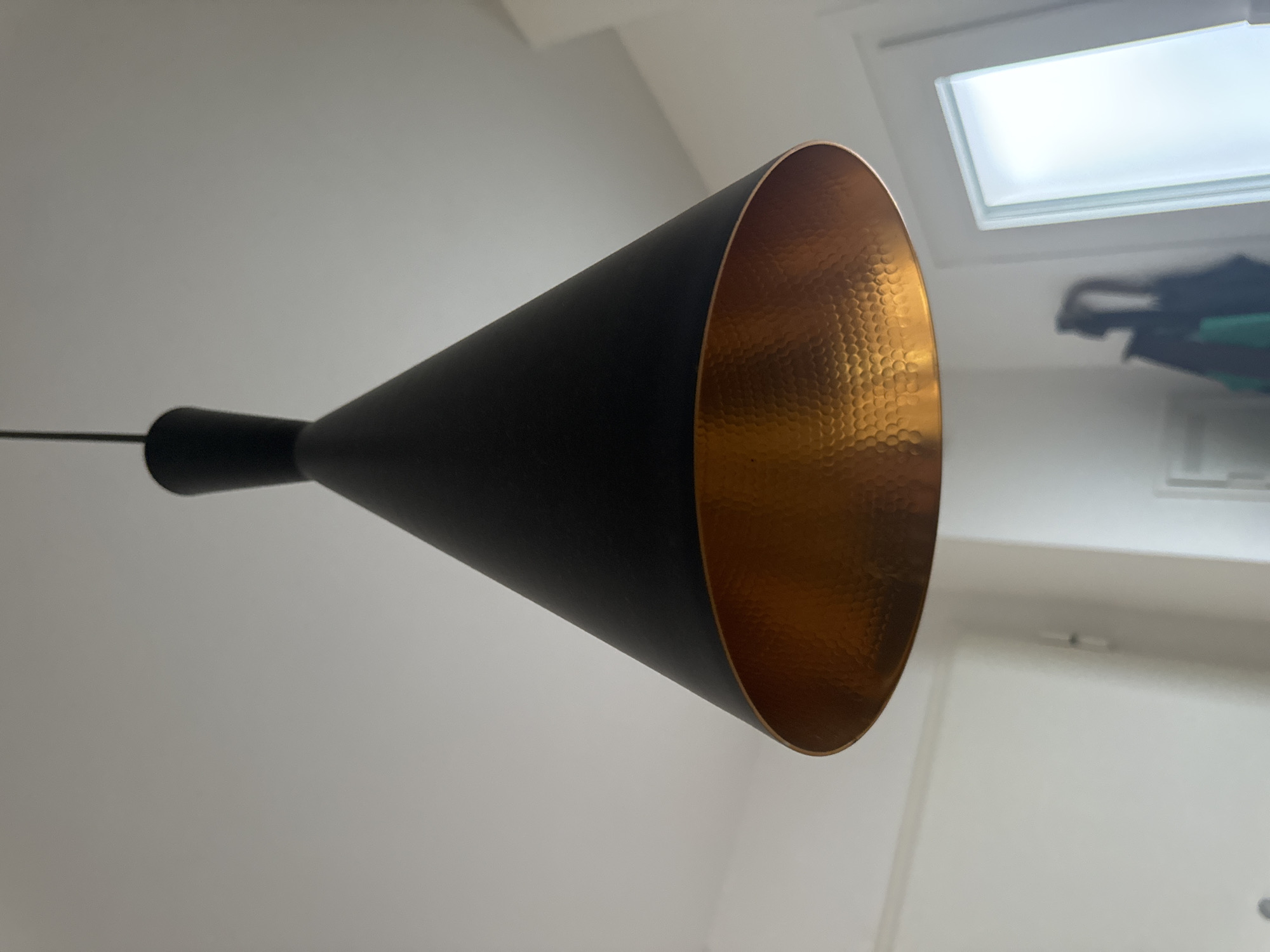 This is a close-up, eye-level shot of a black conical pendant light fixture hanging from a white ceiling. The inside of the fixture is a hammered copper color, reflecting light. The background is a blurry interior scene with white walls and a frosted glass window or door on the right. The location is not specified in the image.