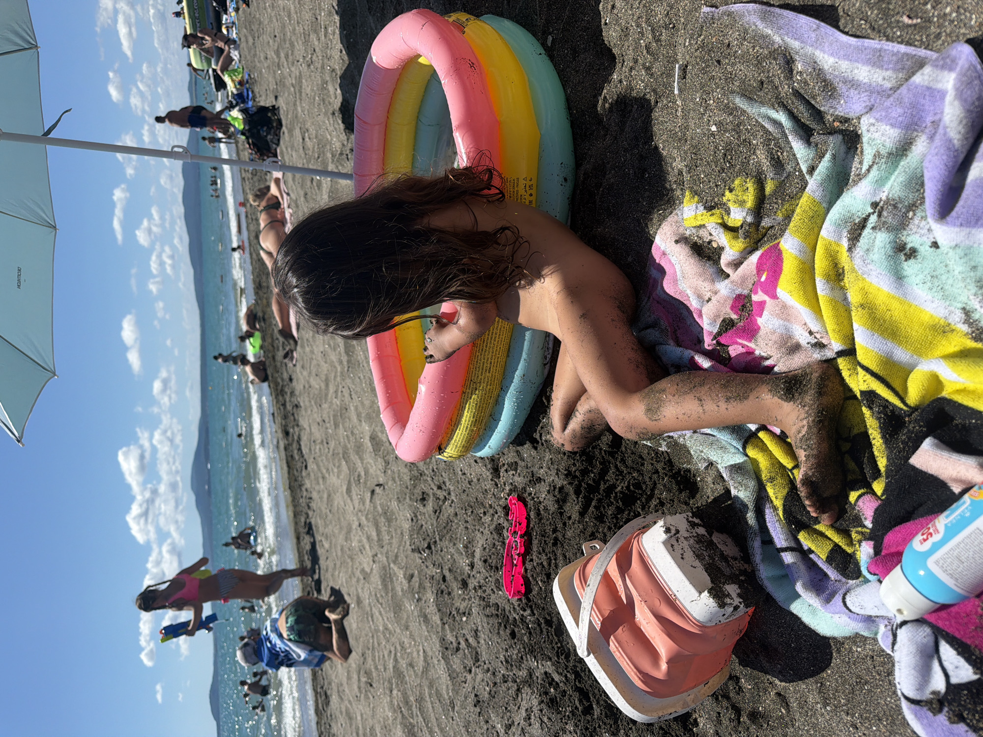 A young girl is sitting on a sandy beach, covered in sand, next to a colorful inflatable pool. The beach is dark, possibly volcanic sand. The girl is looking down at her hands.
In the background, the Black Sea is visible with gentle waves. Several other people are on the beach, some in the water. There are colorful towels and beach accessories scattered on the sand, including a pink toy shovel and a collapsible bucket. A light blue umbrella is partially visible on the left side of the image. The sky is clear and blue, suggesting it is a sunny day.
The scene appears to be a casual day at the beach, likely during the summer. The overall mood seems relaxed and summery. The location is implied to be Burgas, Bulgaria, given the Black Sea. There is some text visible on a blue bottle of sunscreen or lotion near the towels, but it is too blurry to read.