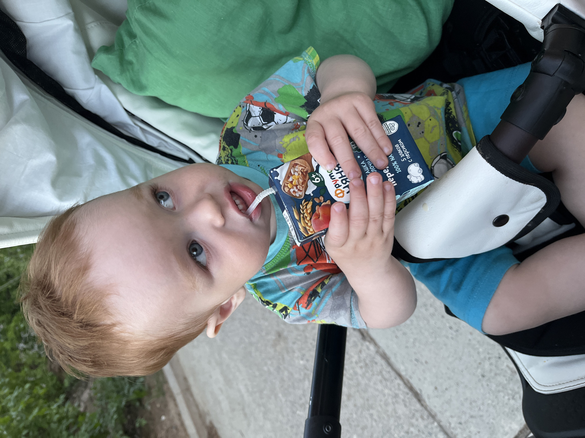 A young child is sitting in a stroller and drinking from a juice box. The child has reddish hair and blue eyes, and is wearing a colorful shirt and blue shorts. The stroller appears to be in a public outdoor space in Chaikovsky, Russia. The area seems to be partially paved.