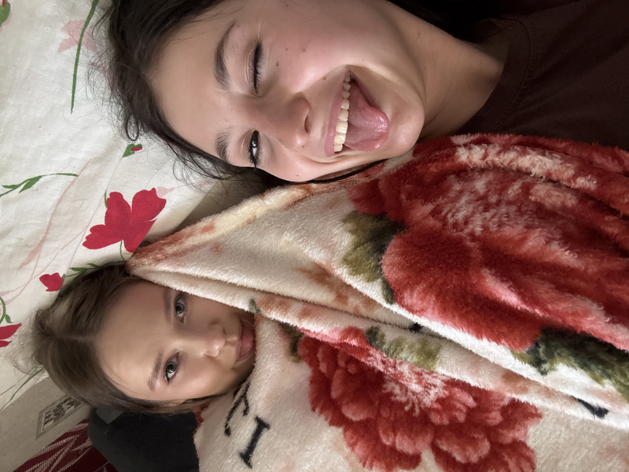 Two young women are lying down next to each other. One is lying on top with her tongue out and smiling. The other is lying beneath a floral blanket, looking at the camera. They are both lying on a patterned bed sheet. There is no indication of the city of Фоки, Russia.