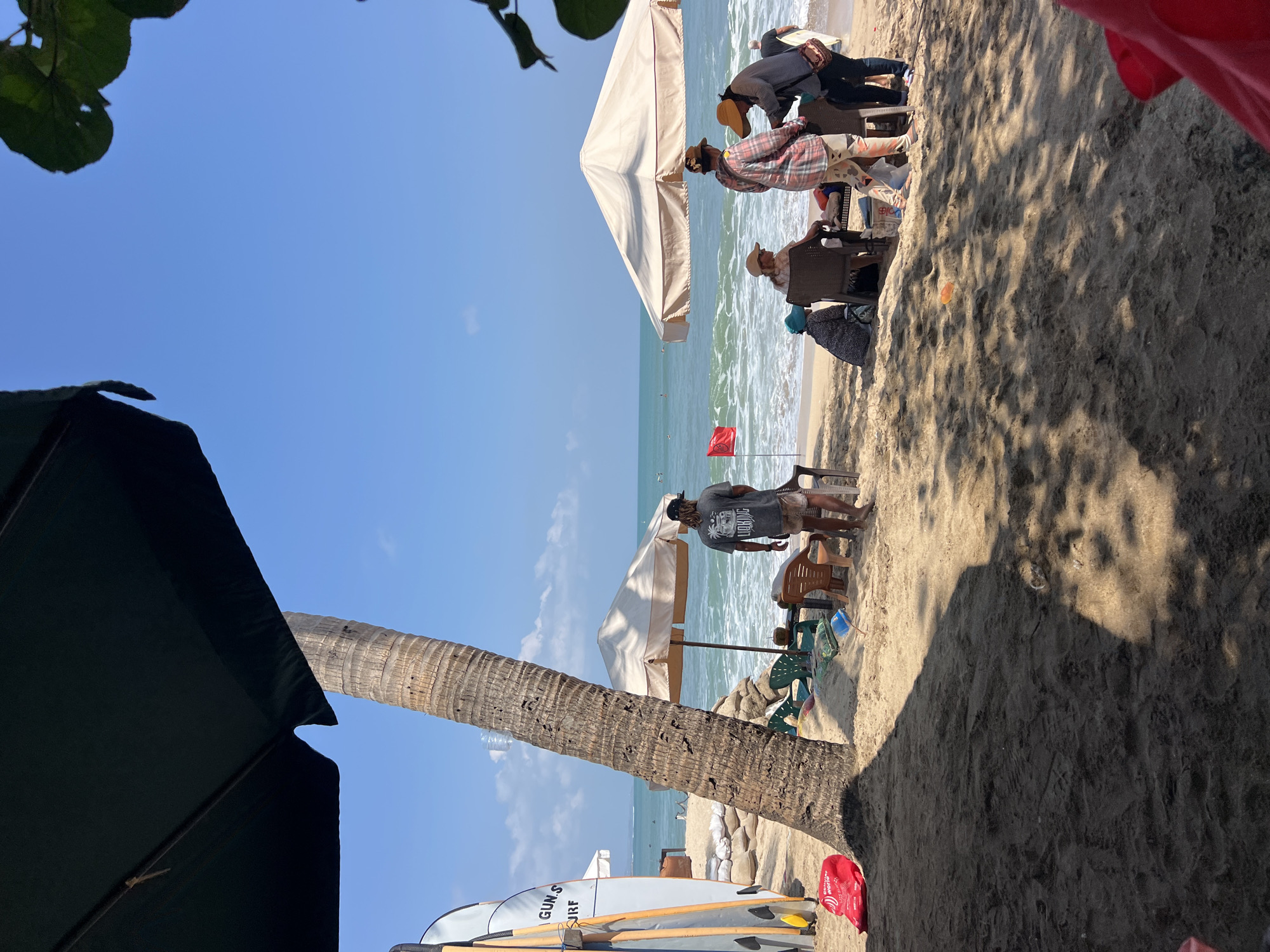 This image depicts a sunny day at Kuta Beach, Indonesia. In the foreground, dark sand is prominent, with shadows cast from an unseen umbrella and foliage. To the left, several surfboards are propped upright. A tall palm tree stands near the center, with its rough bark clearly visible.
Further back on the sand, there are a few beach chairs and umbrellas. A group of people are gathered near a light-colored umbrella. One person, wearing a dark t-shirt and patterned shorts, stands looking out at the ocean. To their right, two individuals are interacting, one wearing a plaid shirt and the other a dark jacket and hat. Another person, wearing a patterned head covering and dress, is seated on the sand to the left of this group.
The ocean is visible in the background, with gentle waves rolling towards the shore. A red flag is planted in the sand near the water's edge. The sky is a clear, bright blue, with a few faint clouds. The scene appears to be a casual beach setting, likely a popular tourist spot.