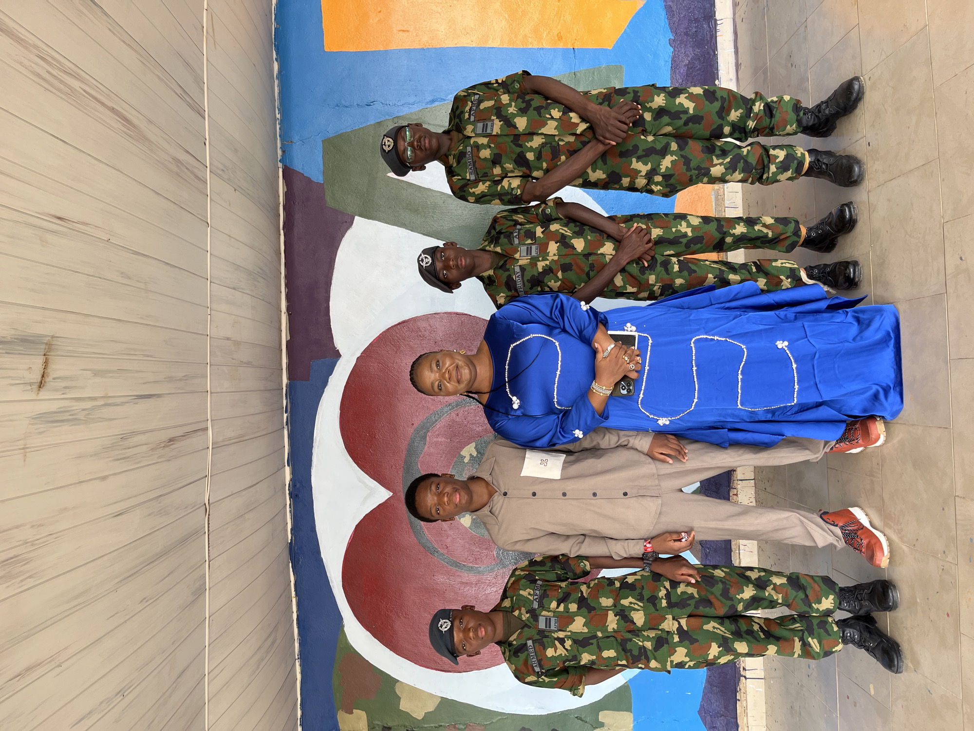 In Bar Jirgi Summa, Nigeria, a group of five individuals stands in front of a colorful mural featuring a large heart. On the far left, a young person in a camouflage military uniform and a black beret looks forward. Next to them, a young male in a tan button-down shirt and matching pants also faces forward. In the center, a woman in a bright blue dress with intricate white beading is holding a smartphone and a book. To her right, another individual in a camouflage military uniform and a black beret stands attentively. Finally, on the far right, a fifth person, also in a camouflage military uniform and a black beret, poses for the photo. The mural behind them is abstract and vibrant, with sections of blue, orange, green, and red. The ground beneath them is tiled.