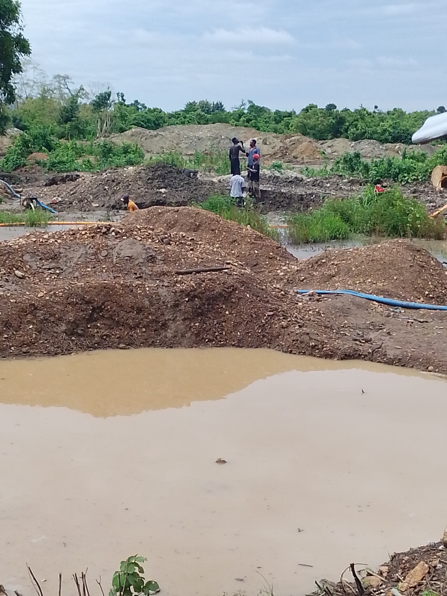 The image depicts an outdoor scene in Otulu, Nigeria, characterized by a muddy, excavated area with piles of soil and debris. In the background, lush green vegetation and trees form a dense backdrop against a cloudy sky. Several individuals are visible within the excavated area, seemingly engaged in work. One person is dressed in a dark garment, another in a red top, and a third in a light-colored shirt. A fourth individual, wearing an orange top, is partially obscured behind a mound of soil. The foreground is dominated by a large, muddy pool of water. A blue hose is seen extending across a pile of gravel in the middle ground. The overall impression is of an active construction or excavation site under overcast conditions.