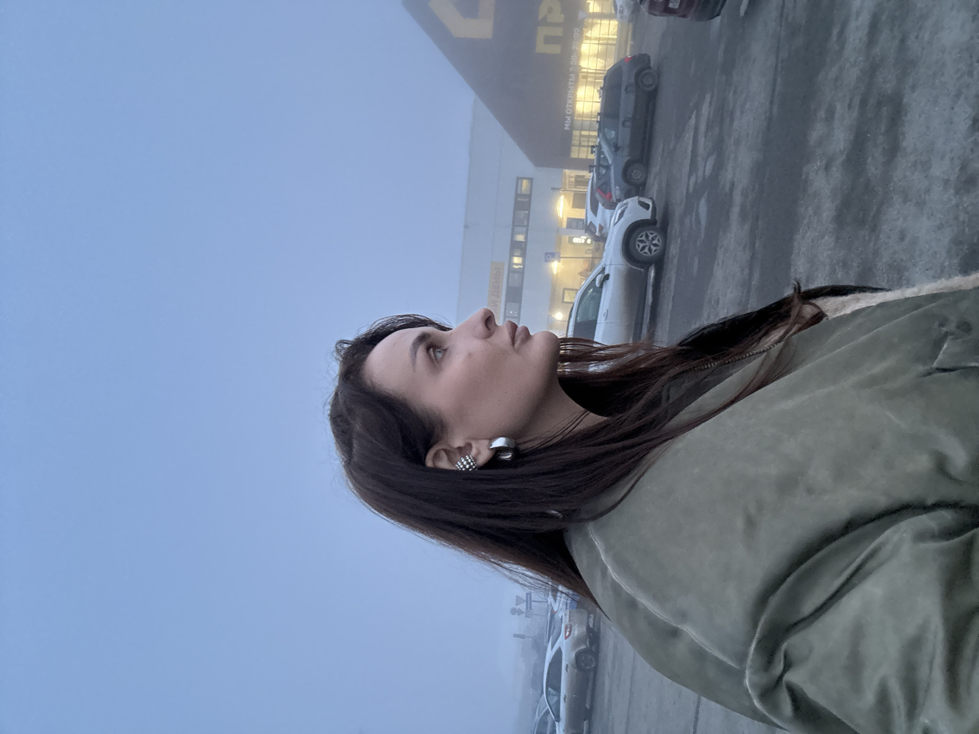 A person with dark hair and fair skin, wearing a green jacket, is looking upwards in an outdoor setting. They are wearing two earrings on the left ear. The background shows a foggy, overcast day, with a building that appears to be a large store or shopping center. Several parked cars are visible, suggesting a parking lot in front of the building. The building has visible signage in what appears to be Russian Cyrillic script, indicating "Мы открыты 7.00-22.00". Based on this and the foggy, overcast environment, the location appears to be Izhevsk, Russia.