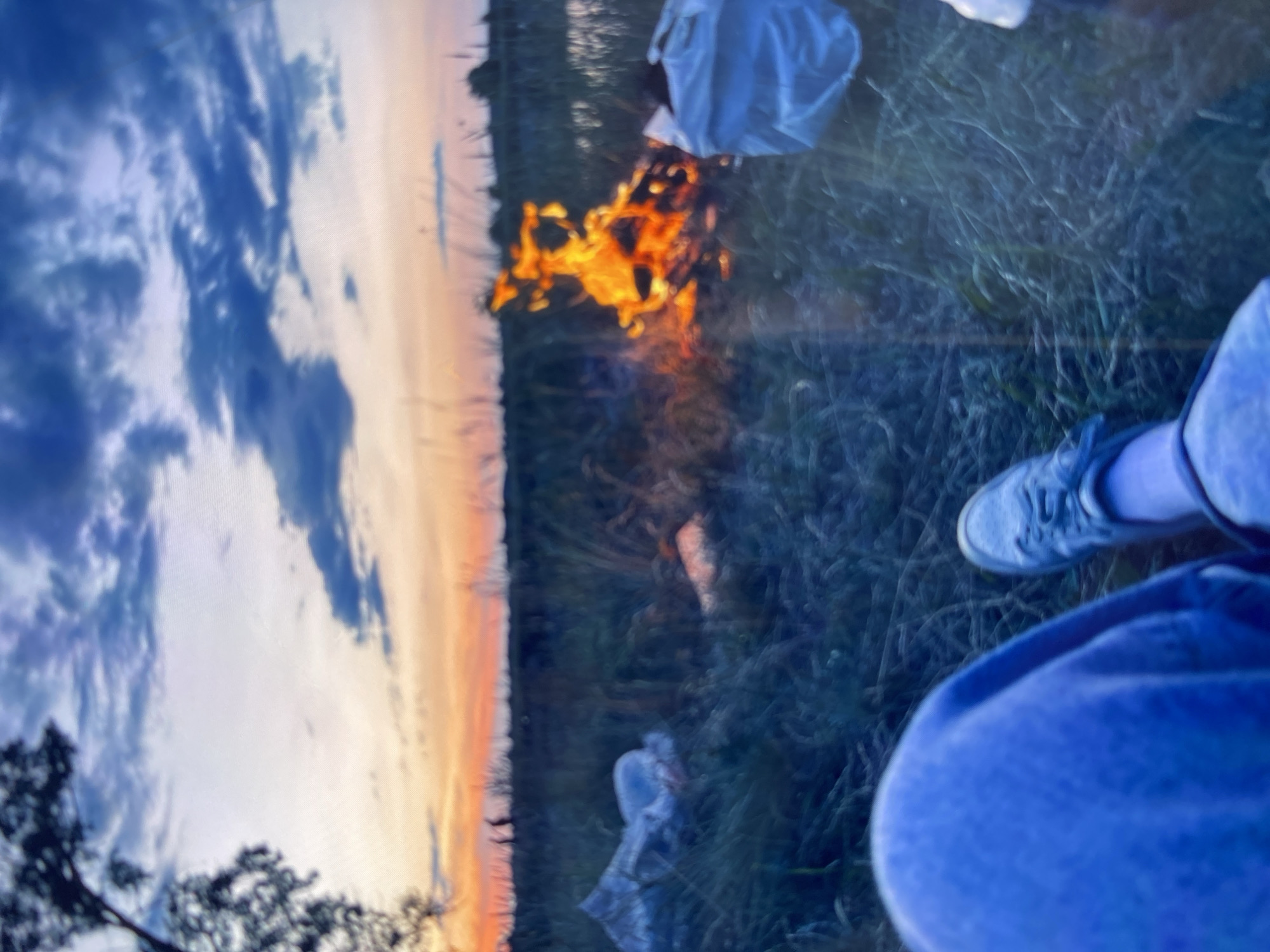 A low-angle, blurry, medium shot shows a person's legs and feet in blue pants and grey sneakers, sitting on grass by a campfire at dusk. The fire, in the center of the frame, burns brightly with orange flames and emits smoke. To the left of the fire, a white plastic bag is visible among the grass, and to the right, another white plastic bag is partially obscured by the foreground. The background features a field with tall grass, a distant treeline, and a sky filled with dramatic clouds in shades of blue, purple, and orange, indicating either sunrise or sunset. The overall scene conveys a sense of an outdoor gathering or camping.