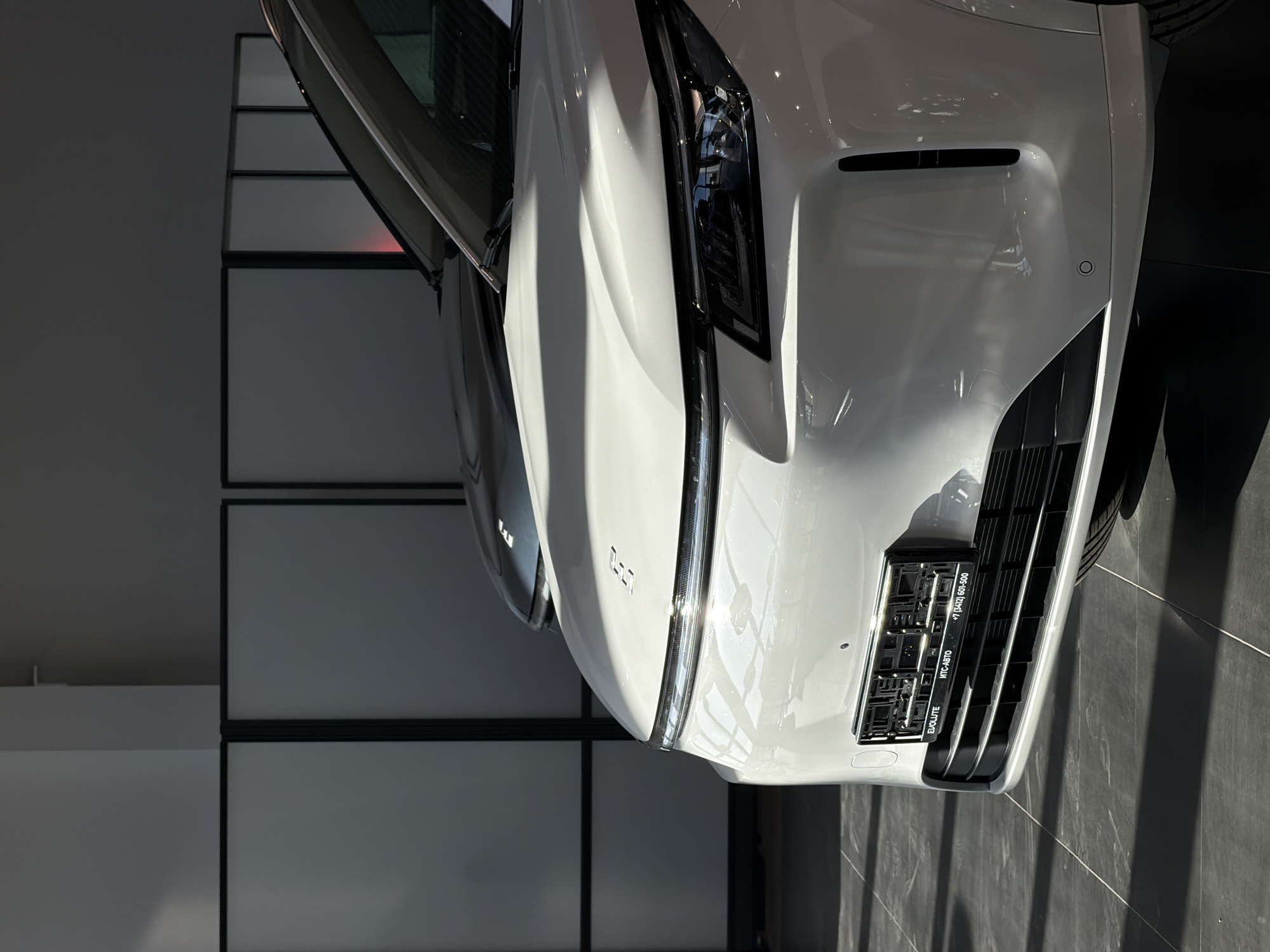 A close-up, angled, indoor shot shows the front of a white car with the driver's side door slightly ajar, positioned on a gray tile floor. The car's front grill is visible, with a license plate frame that includes the words "EVOLUTE" and "+7 (3412) 601-500". Behind the car, a wall composed of gray rectangular panels outlined in black is visible. The location is likely a car dealership or showroom. The city of Izhevsk, Russia is mentioned in the details of the phone number +7 (3412) which is the telephone code for Izhevsk.