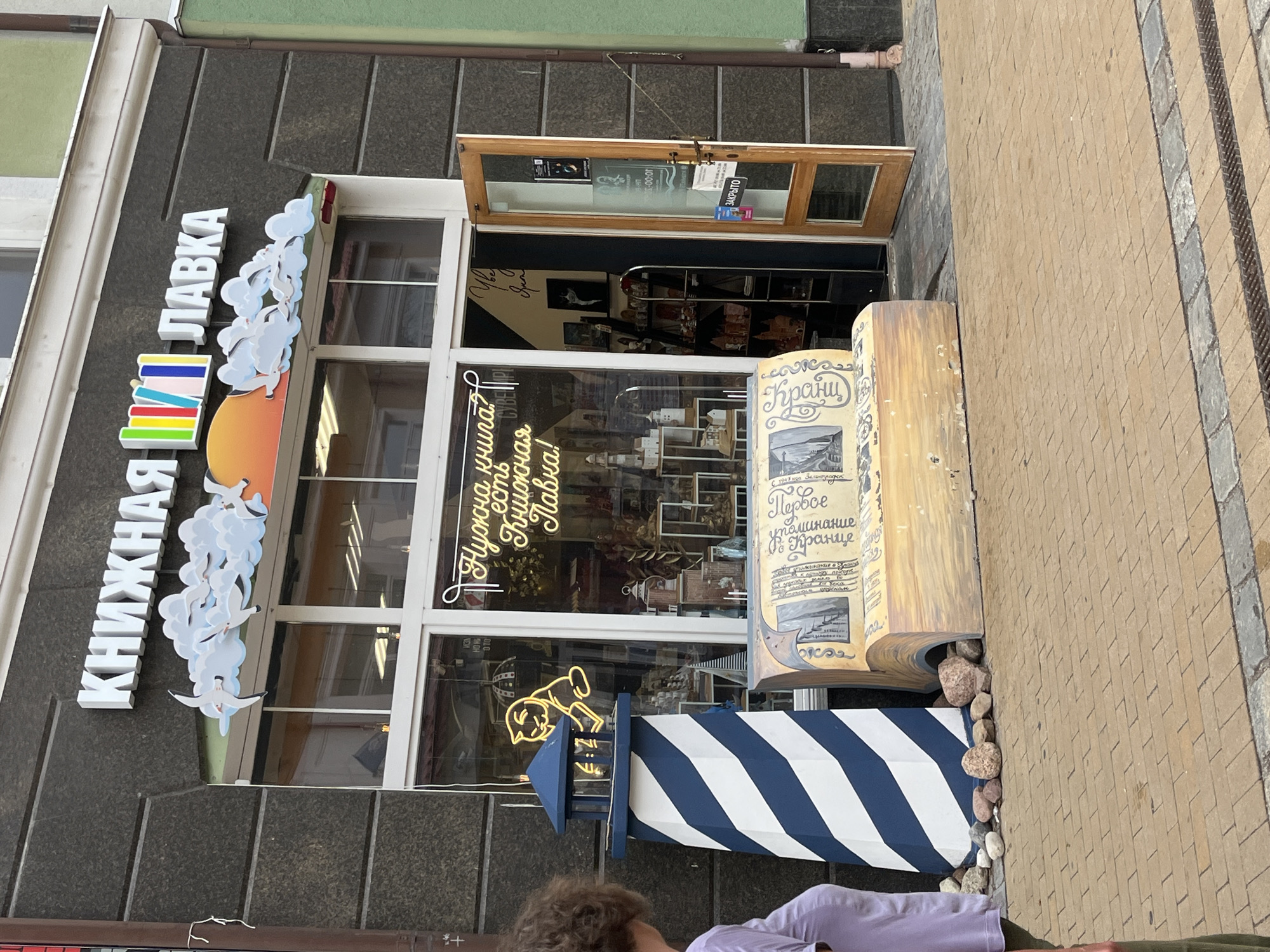 This image depicts the exterior of a bookstore named "КНИЖНАЯ ЛАВКА" (Knizhnaya Lavka) located in Zelenogradsk, Russia. The store's facade features a large window display and a prominent sign above. Decorations include stylized clouds with seagulls and a colorful representation of books. In front of the store, a lighthouse-shaped object and a bench designed to resemble a stack of books are visible. A person, partially cropped on the left, stands facing away from the camera. The ground is paved with brickwork. The store appears to be closed, as indicated by a "ЗАКРЫТО" (Closed) sign on the glass door.