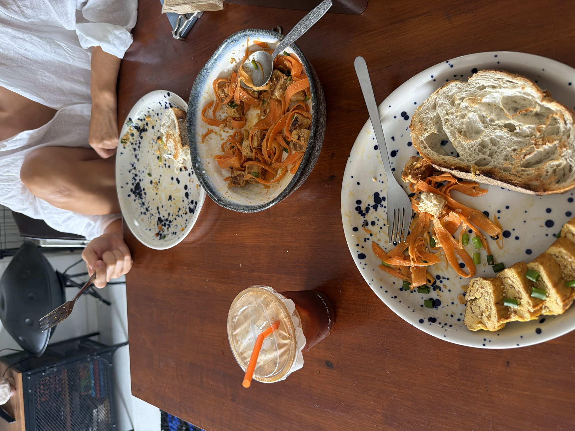 The image provides an overhead view of a person enjoying a meal at a dark wooden table in an indoor setting, likely a cafe or restaurant in Chang Phueak, Thailand. A person, visible from the torso downwards, is wearing a loose-fitting white top. Their right hand is seen holding a fork, indicating they are in the process of eating.
On the table, there are three ceramic plates and one bowl, each adorned with a distinctive white base and blue/black splatter patterns. One plate to the right holds a thick slice of sourdough bread, several pieces of sliced tamagoyaki (rolled omelet) garnished with chopped green onions, and some orange, ribbon-cut carrots. A central bowl contains a dish of orange carrot ribbons, cubes of what appears to be tofu or tempeh, and green onions, served with a decorative metal spoon. Another plate to the left is mostly cleared, showing only crumbs and sauce residue.
A clear plastic cup with a lid and an orange straw sits on the table, containing a dark iced beverage. The text "N/ACC" is faintly visible and repeated on the side of the cup. The food items show signs of having been partially consumed.
In the background, elements of the room include a black chair or stool, a metal mesh cabinet with various items stored inside, and a dark, rounded object resembling a hang drum or speaker. The scene suggests a relaxed daytime meal or snack.