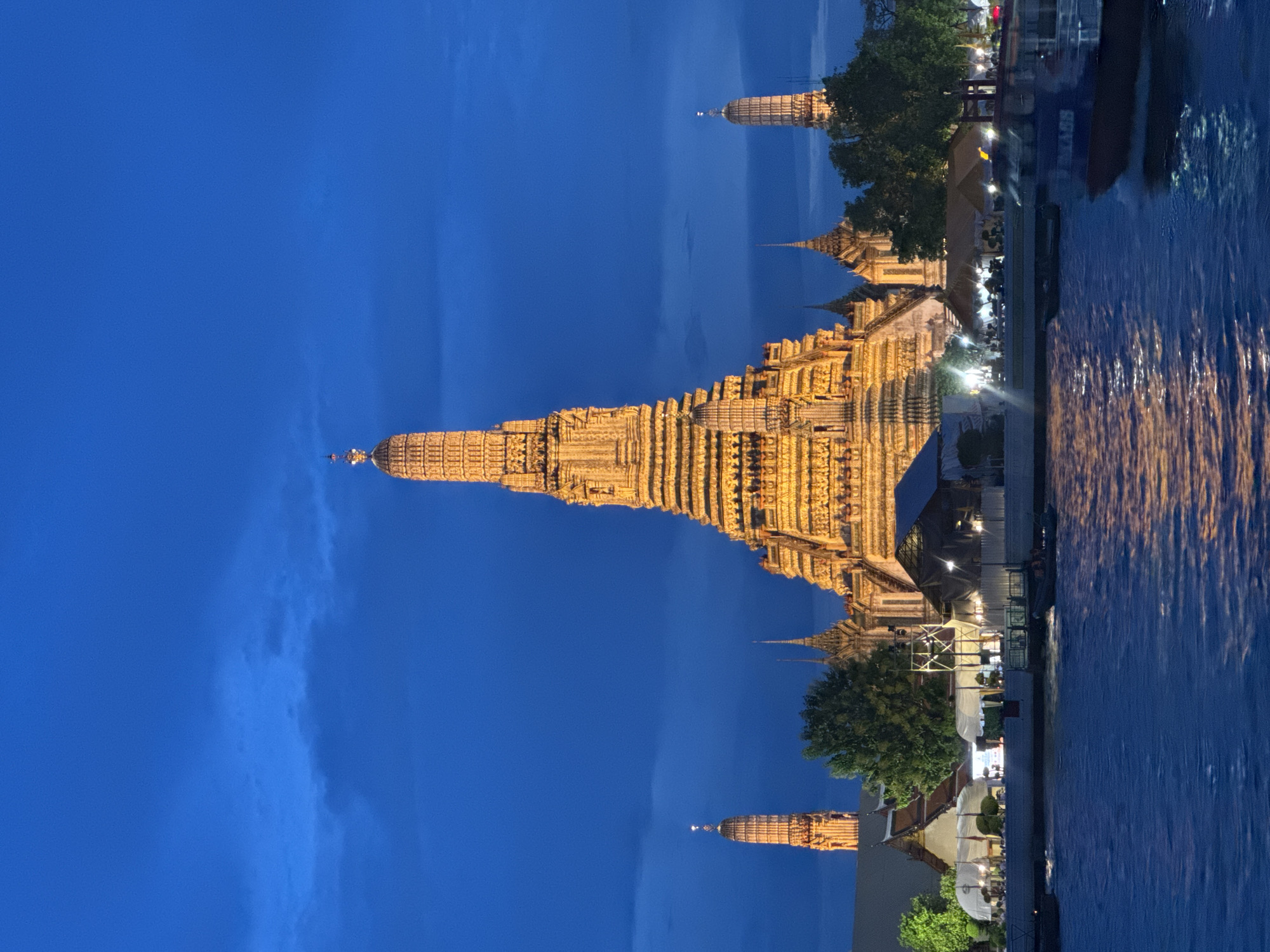 This image depicts the Wat Arun temple in Bangkok, Thailand, illuminated at dusk. The temple's prominent prang, a towering spire, is the main subject, glowing with warm light against a deep blue sky. Surrounding structures of the temple complex are also visible, as are trees and what appears to be a body of water in the foreground, possibly the Chao Phraya River, reflecting the temple's light. Several smaller spires of the temple are also visible. There is a hint of people and activity along the riverbank, with lights suggesting businesses or restaurants. The overall mood is serene and majestic, highlighting the beauty of the temple at twilight. There is no visible text in the image.