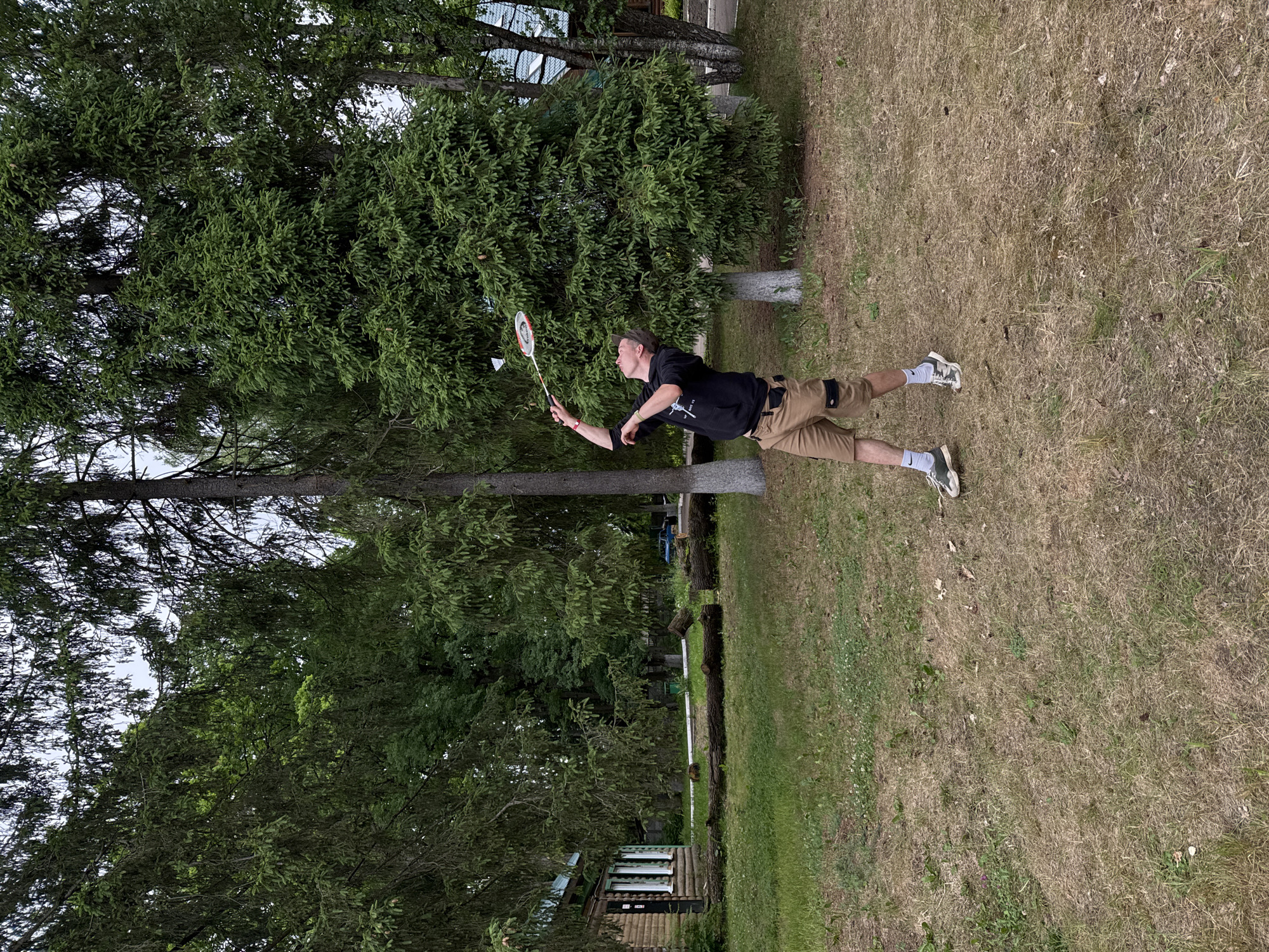 A person is playing badminton outdoors on a grassy area with trees in the background. The person is a male wearing a black t-shirt, khaki shorts, white socks, and athletic shoes. He is mid-air, lunging to hit a shuttlecock with a badminton racket. The scene appears to be set in a park or a backyard with a mix of green grass and dry, patchy areas. Tall evergreen trees and some deciduous trees form a dense backdrop. There are also some structures visible in the background, including what looks like a small wooden building. The lighting suggests it is daytime, possibly overcast, as there are no strong shadows. The overall mood is active and recreational.