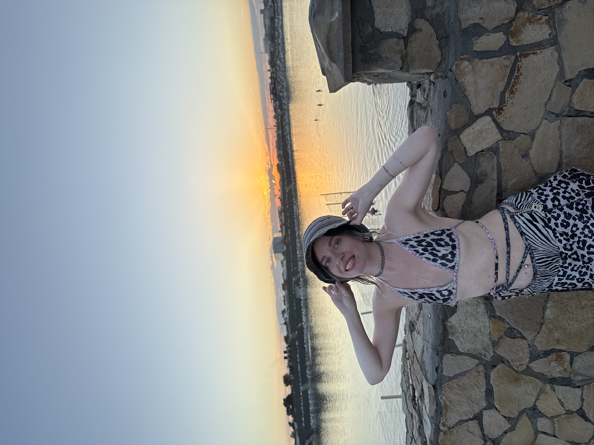 A young woman stands on a stone wall overlooking a body of water at sunset. She is wearing a bucket hat, a leopard-print bikini top, and a patterned skirt with zebra and leopard print. Her hands are resting on her hat, and she is smiling at the camera. The sun is setting over the water, casting an orange glow on the sky and water. In the background, there are buildings and trees on the horizon. The scene appears to be a public area, possibly a park or promenade, in Sol-Iletsk, Russia, given the presence of the water and the general landscape, although specific landmarks are not identifiable in this image.