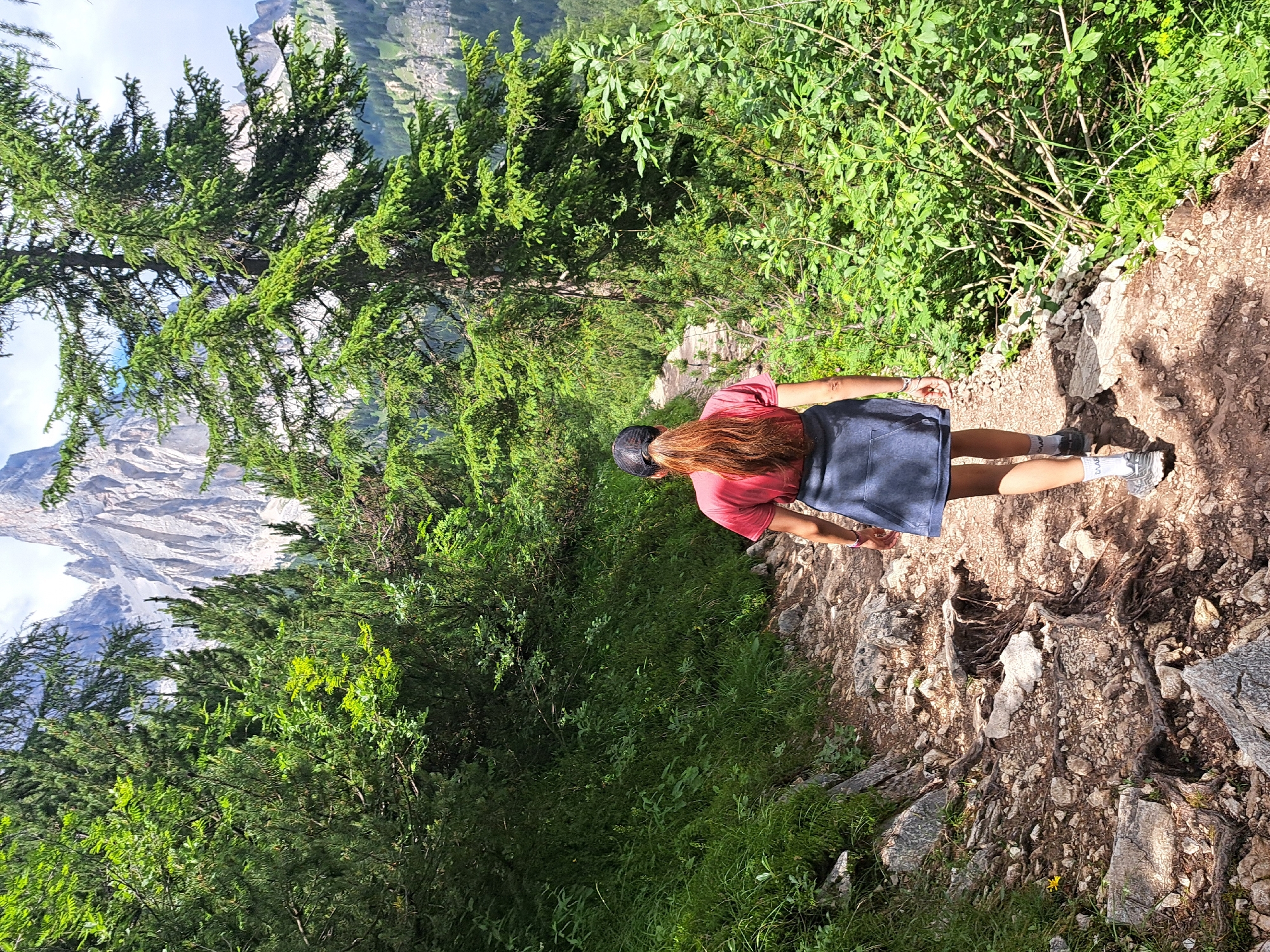 The image shows a person from the back, wearing a red t-shirt, a dark skirt, a black cap, and grey sneakers, walking on a dirt path. The person has long, reddish-brown hair. The path is surrounded by lush green vegetation, including trees and bushes. In the background, a majestic mountain range with rocky peaks is visible, partially obscured by trees and foliage. The lighting suggests it is daytime, likely sunny, with shadows cast on the path. The overall scene depicts a person hiking or walking in a natural, mountainous environment, possibly during a summer day given the green foliage and the clear sky. The image is oriented vertically. There is no visible text.