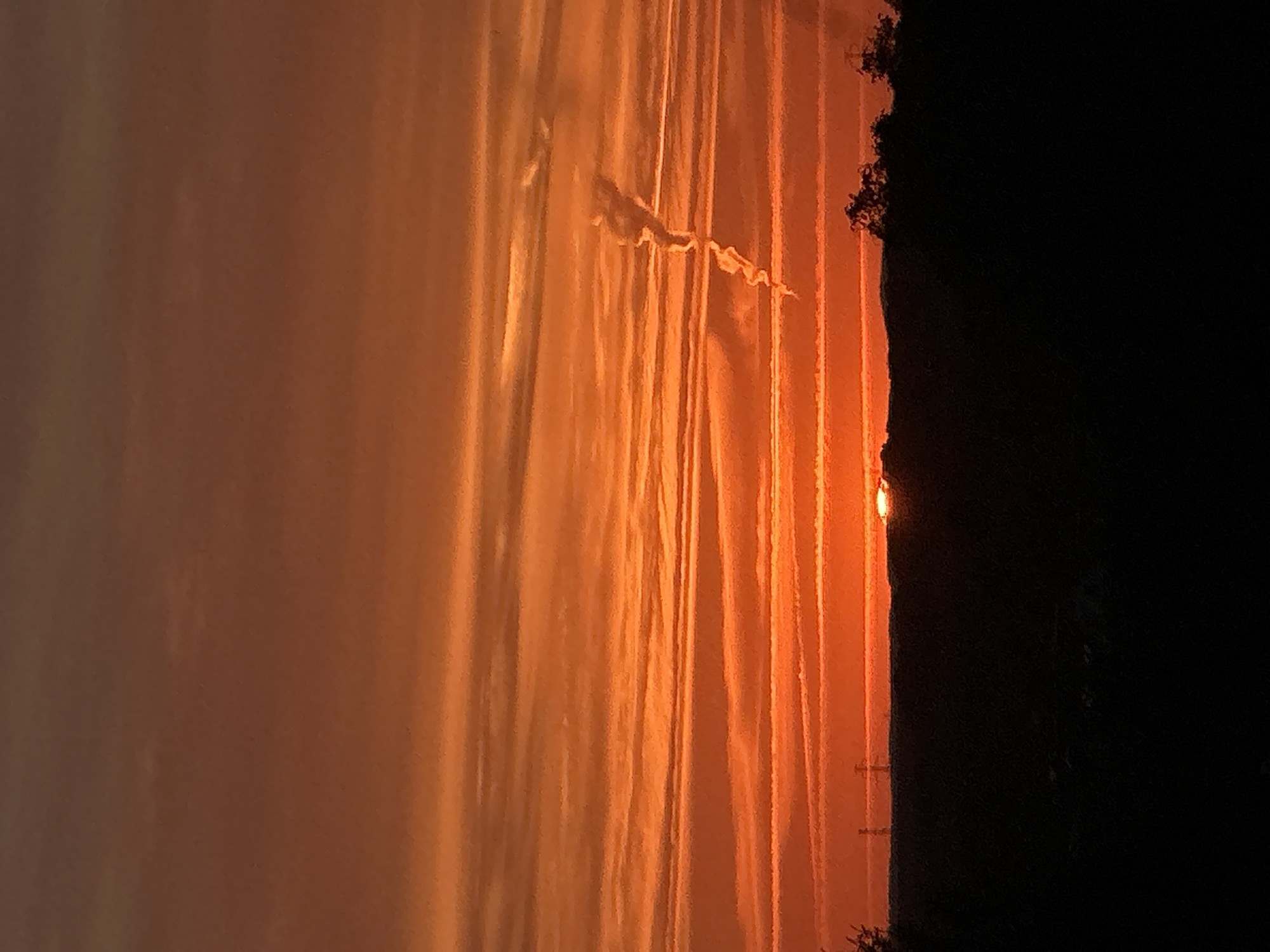 The image captures a dramatic sunset over the city of Świdnica, Poland. The sky is ablaze with vibrant hues of orange and red, streaked with wispy clouds that form horizontal lines across the frame. A single, elongated contrail trails downwards from the upper right, adding a dynamic element to the celestial display. The sun, a fiery orb, is partially obscured by the dark silhouette of the horizon. This silhouette encompasses a faint outline of power lines on the left and a cluster of trees on the right, indicating an elevated landscape. The foreground is consumed by deep shadow, obscuring any details of the ground or immediate surroundings.
