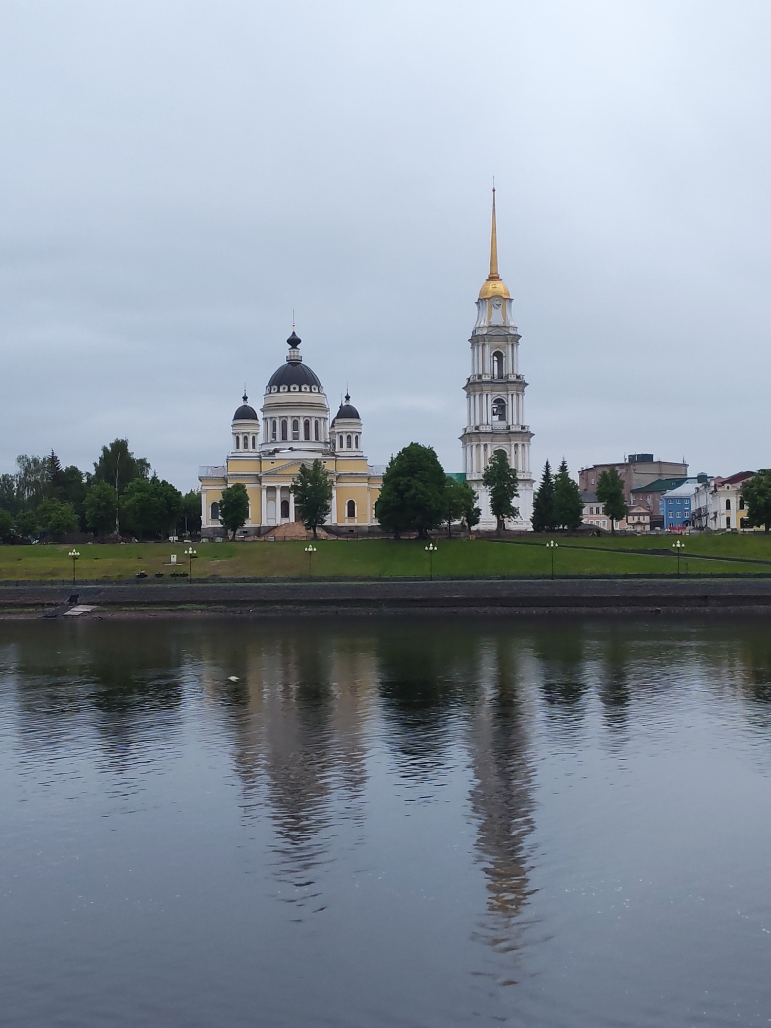 The image depicts a cityscape in Bol'shie Mkhi, Russia, dominated by a large yellow and white cathedral with multiple black domes, and a tall, white bell tower with a golden spire. Both structures are reflected in the calm water in the foreground, likely a river or large body of water. The buildings are situated on a grassy embankment lined with lampposts and trees. Beyond the cathedral and bell tower are other buildings of varying heights and colors. The sky is overcast. A small, dark figure can be seen near the water's edge on the left.