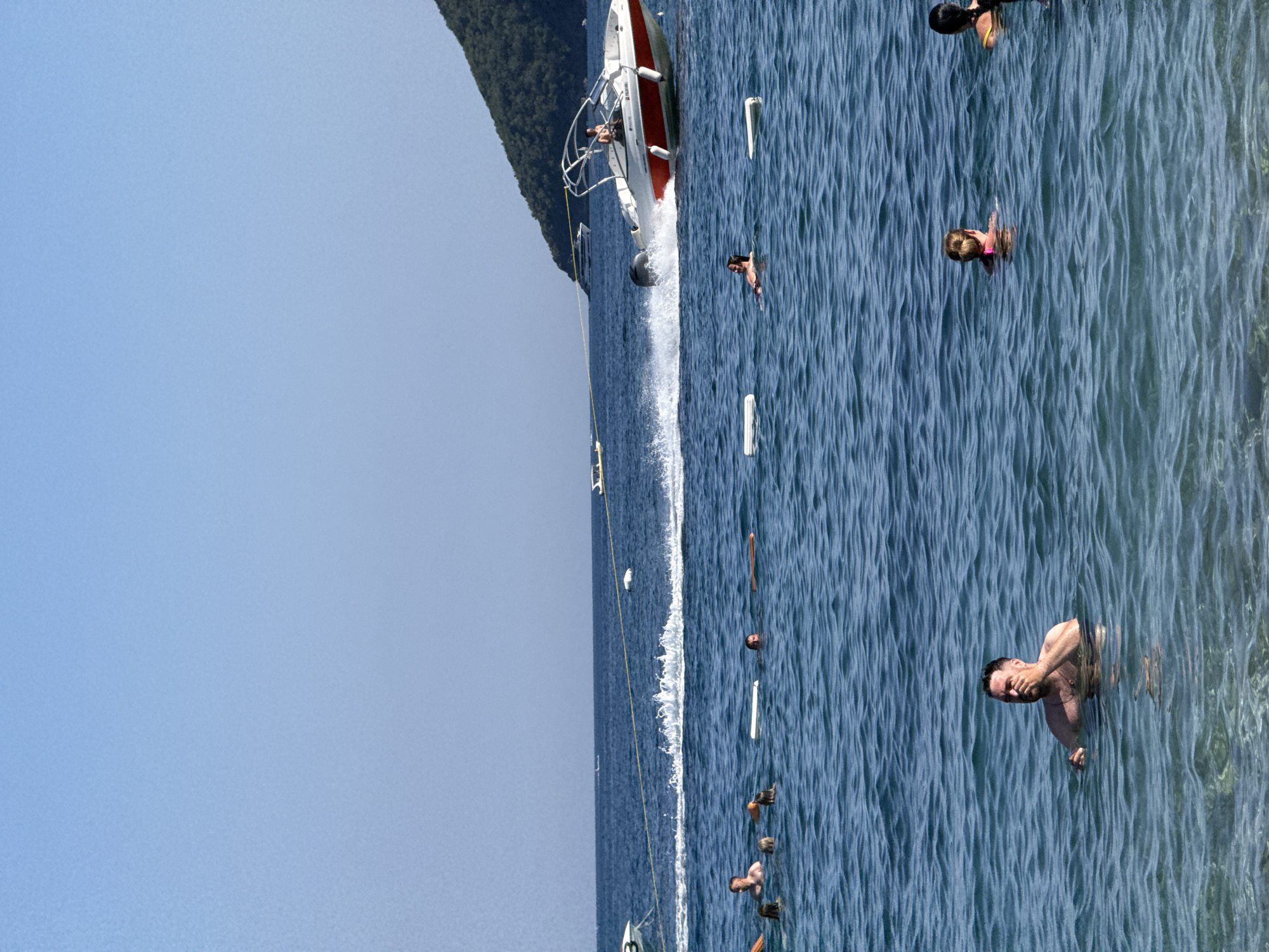 A speedboat is towing a yellow rope across the blue ocean, creating a wake. Several people are swimming and floating in the water. In the foreground, a man is swimming and touching his face. Further back, other swimmers are visible, some with inflatable buoys. A green, tree-covered hill is in the background. The sky is clear and blue. This scene is likely at a beach in Budva, Montenegro, given the hilly coastline and the typical recreational activities observed.