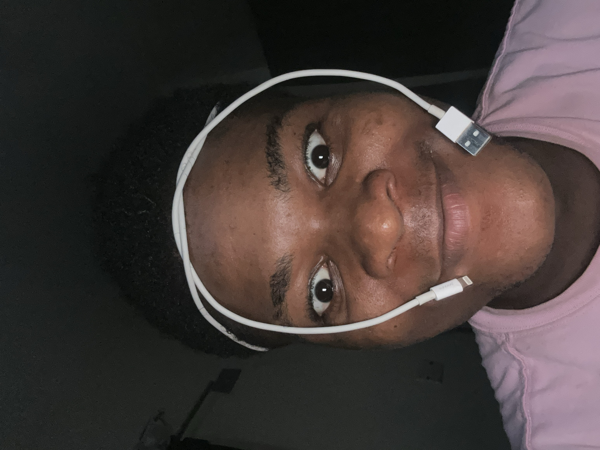 A medium close-up selfie shows a young Black person wearing a pink t-shirt and a white charging cable looped around their head. The cable connects to a lightning port on the left side of their face and a USB-A port on the right side, near their cheek. The person's eyes are wide open, and they are smiling. The background is dark, suggesting an indoor setting at night. The image was captured in Ihiagwa, Nigeria.